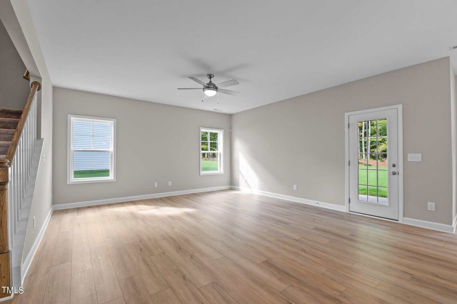 Spacious empty living room with light wood flooring, beige walls, two windows, a ceiling fan, and a glass door leading to a green backyard. Carpeted stairs are visible on the left side.