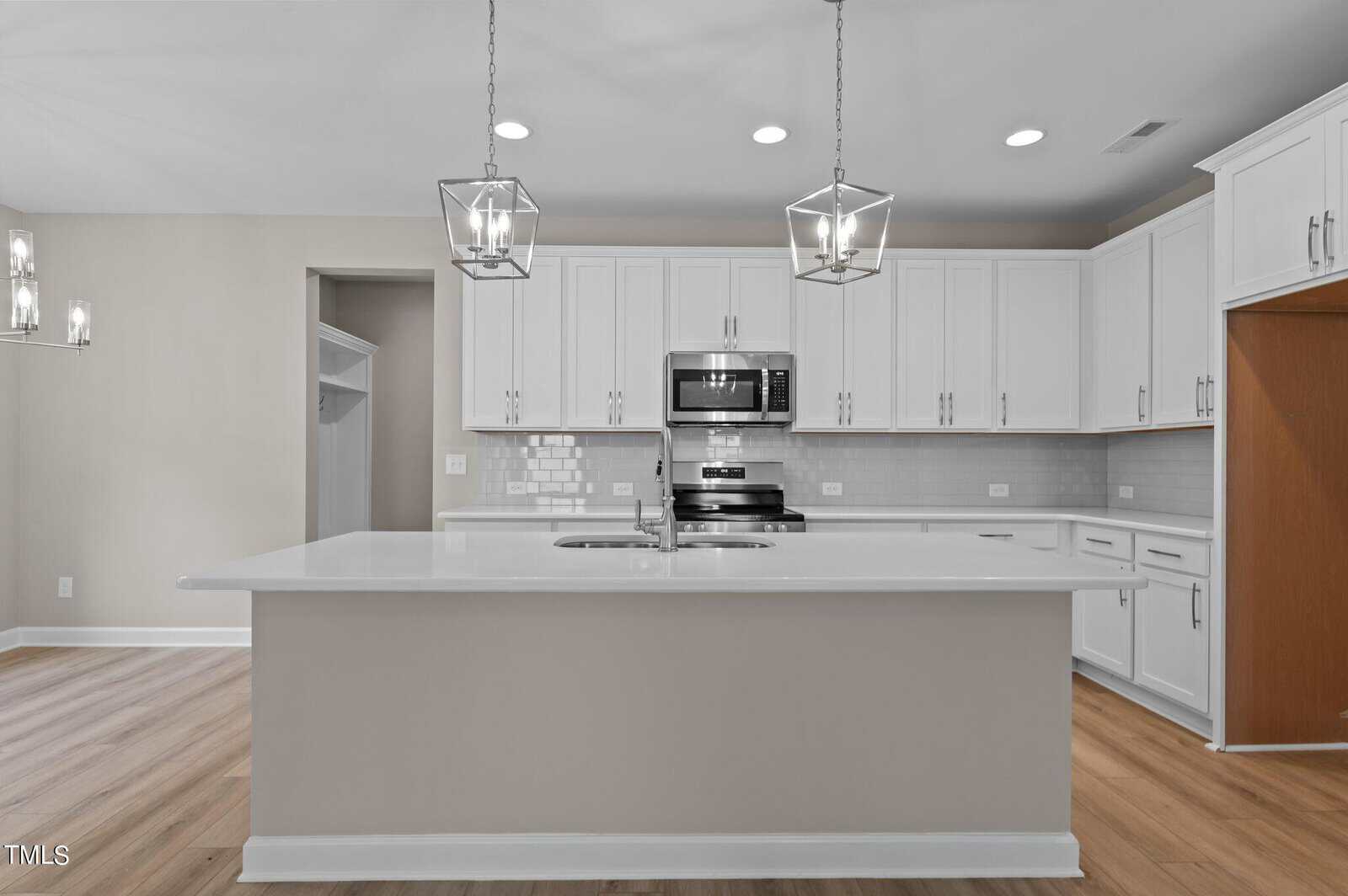 Modern kitchen with white cabinets, stainless steel appliances, white subway tile backsplash, and a large island with a double sink. Two pendant lights hang over the island, and wood flooring is visible throughout.