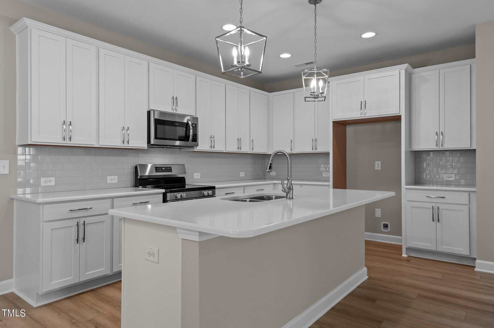Modern kitchen with white cabinets, stainless steel appliances, a large island with a sink, subway tile backsplash, pendant lights, and wood-style flooring. The room is well-lit and has a neutral color scheme.