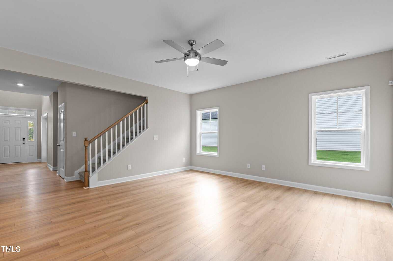 Bright, empty living room with light wood floors, beige walls, two windows, a ceiling fan, and a staircase leading upstairs. Entryway visible to the left with a white front door.