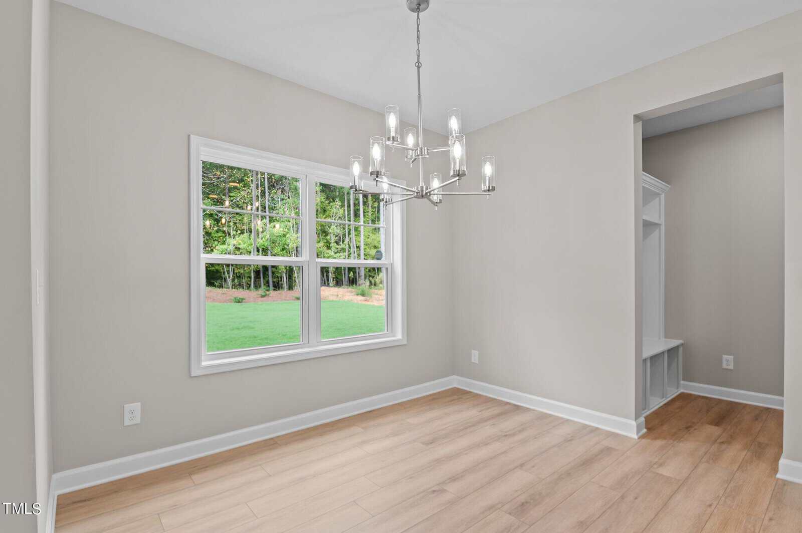 A bright, empty room with light wood floors, beige walls, a modern chandelier, a large window overlooking a green lawn, and an open entry to a built-in bench with storage.
