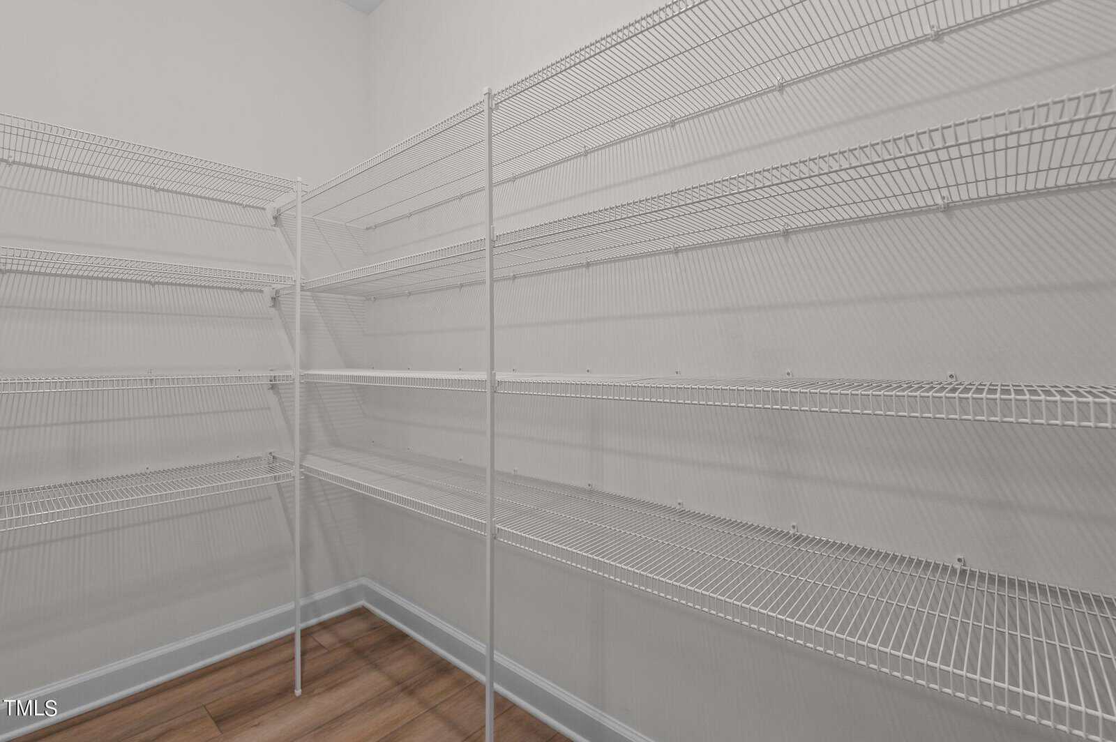 Empty walk-in pantry with multiple rows of white wire shelving on two walls, set against light gray walls and a brown wood floor.