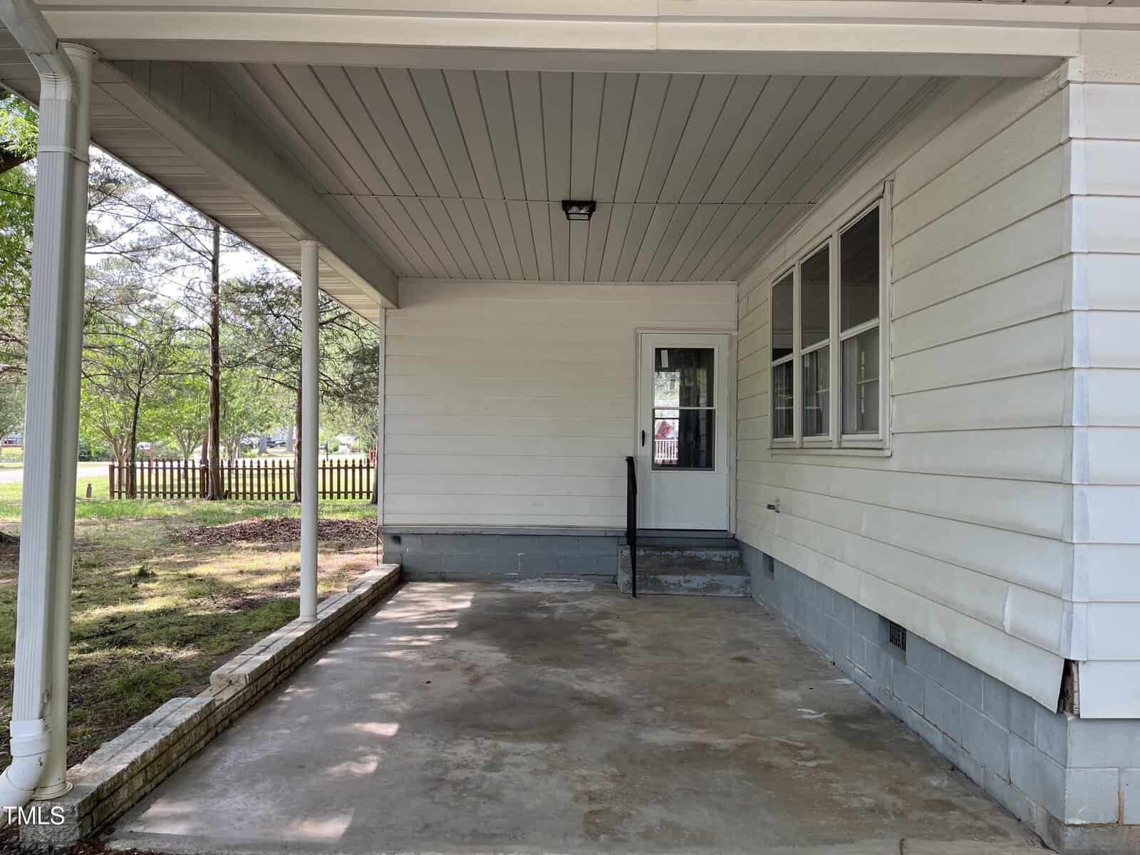 A covered porch with a concrete floor attached to a white house. There's a door with a window and several other windows on the right. A wooden fence and trees are visible in the background.