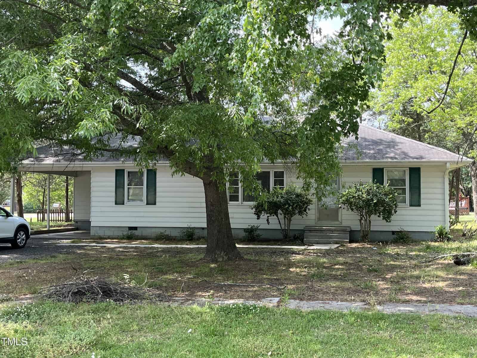 A small, single-story house located at 306 W C Street in Butner features white siding and green shutters, surrounded by trees and lush grass. A large tree shades the front yard, with a driveway leading to a carport on the left side of the home.