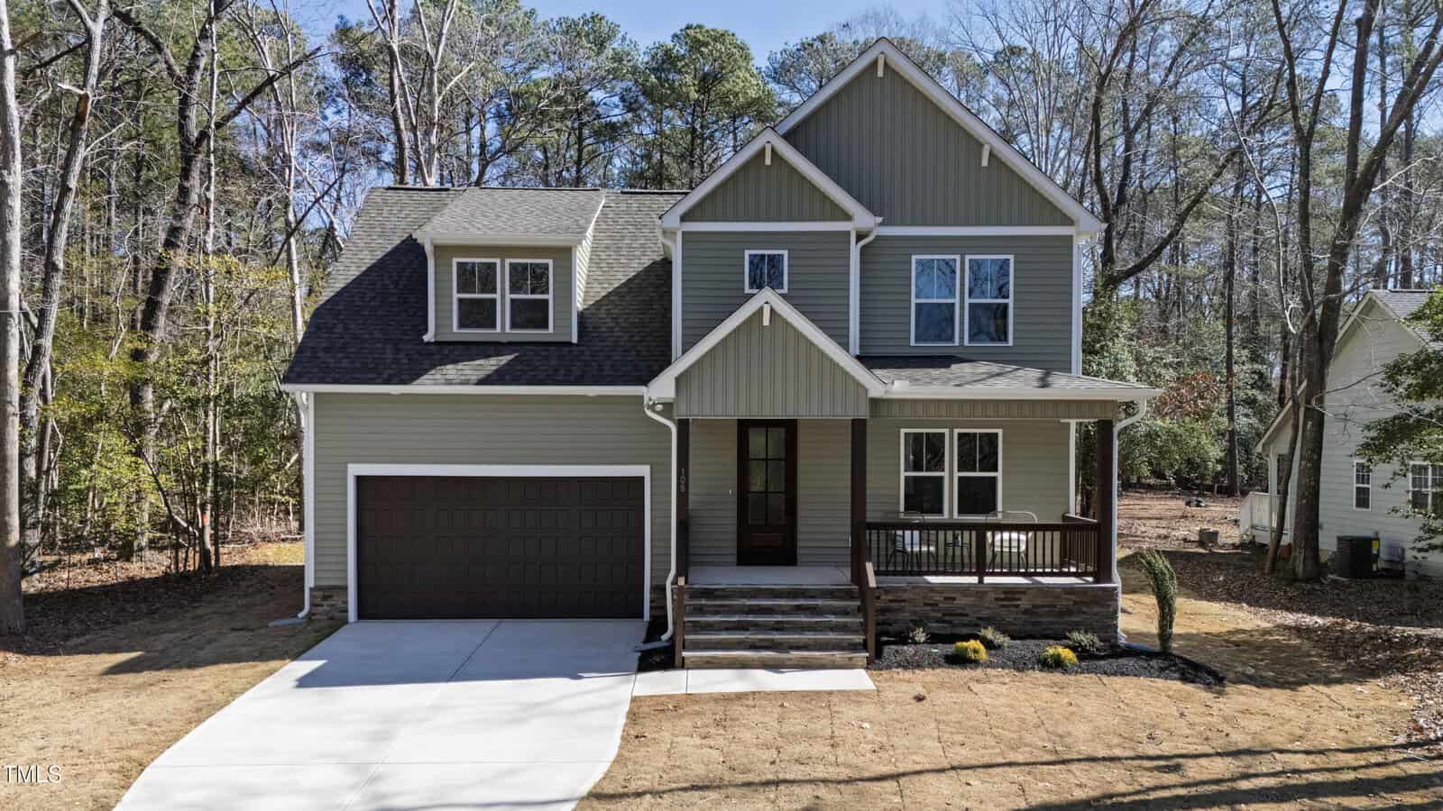 A two-story modern house with gray siding, white trim, and a dark brown garage door, surrounded by trees. The home features a front porch with steps and a newly paved driveway.