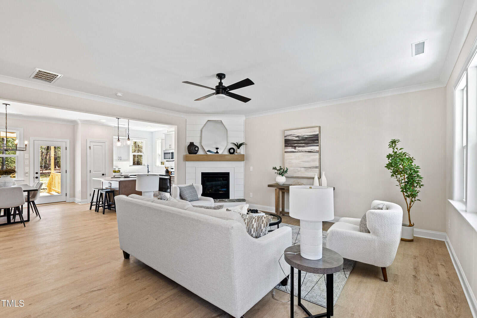 A bright, modern living room with light-colored furniture, a round coffee table, and a fireplace. The space opens to a kitchen with bar stools, and large windows provide natural light. A ceiling fan is centered above.