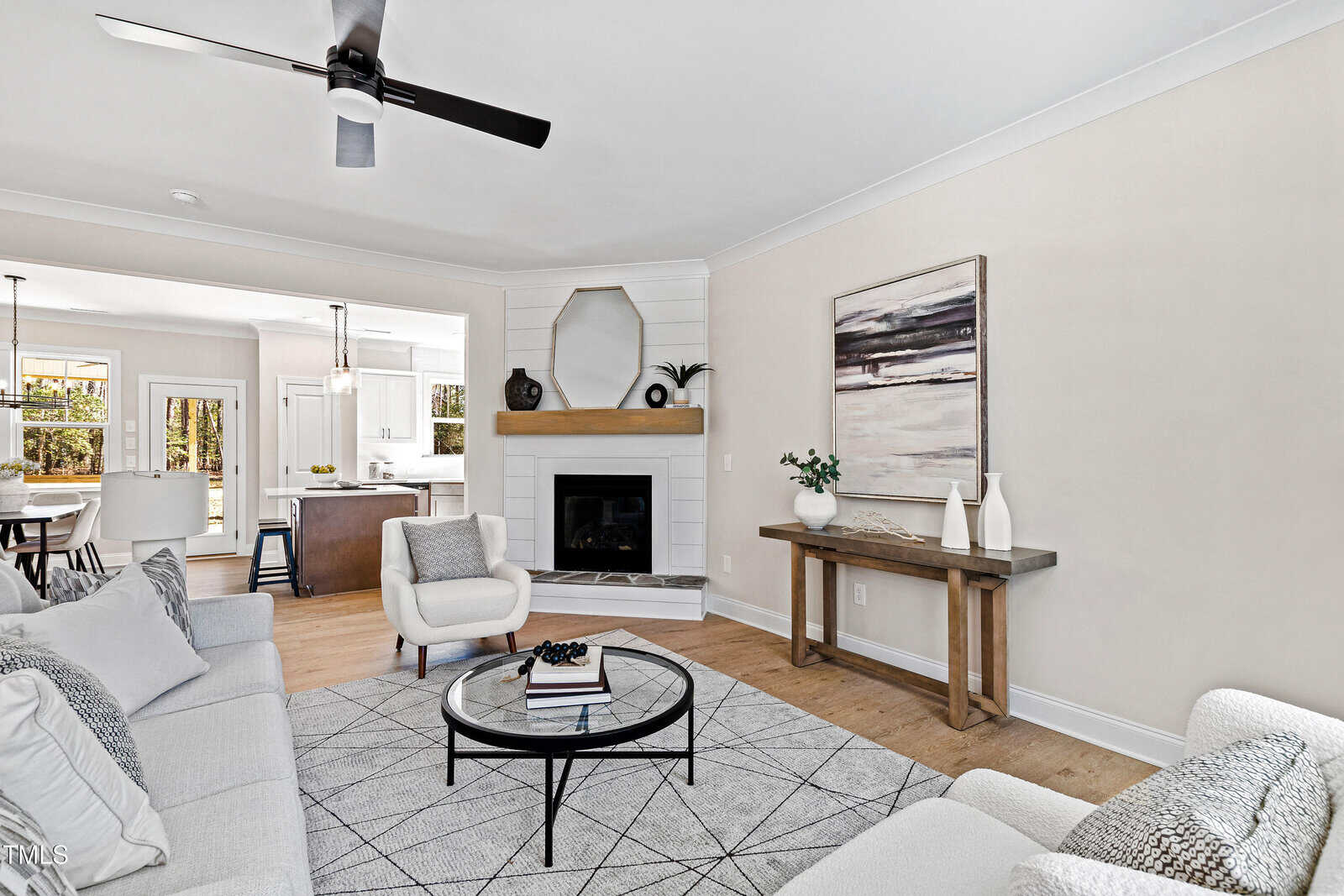 A modern living room with neutral tones features a gray sofa, armchairs, a round patterned rug, and a fireplace with a wooden mantel. Large abstract art and decorative vases sit on a console table, and the space opens to a kitchen and dining area.