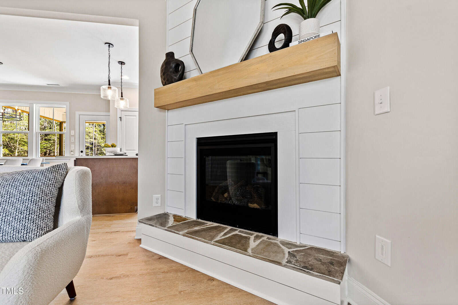 A modern living room with a white shiplap fireplace, a wooden mantel, decorative vases, and a mirror. The hearth has a stone surface, and a kitchen with pendant lights is visible in the background.