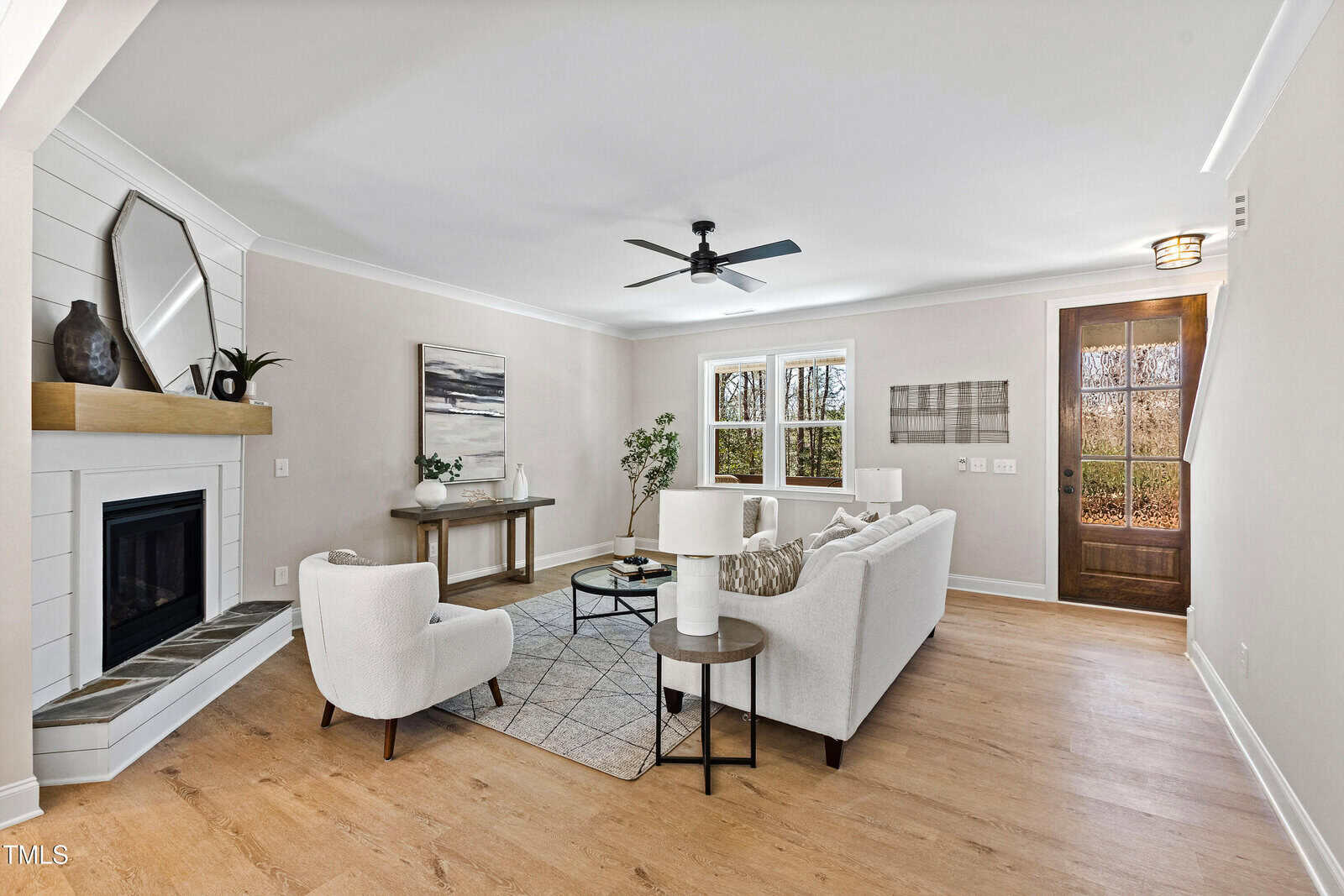 Bright, modern living room with light wood floors, white walls, a white sofa, armchair, round coffee table, wall art, indoor plants, a black ceiling fan, fireplace with a mantel, and large windows near a wooden front door.