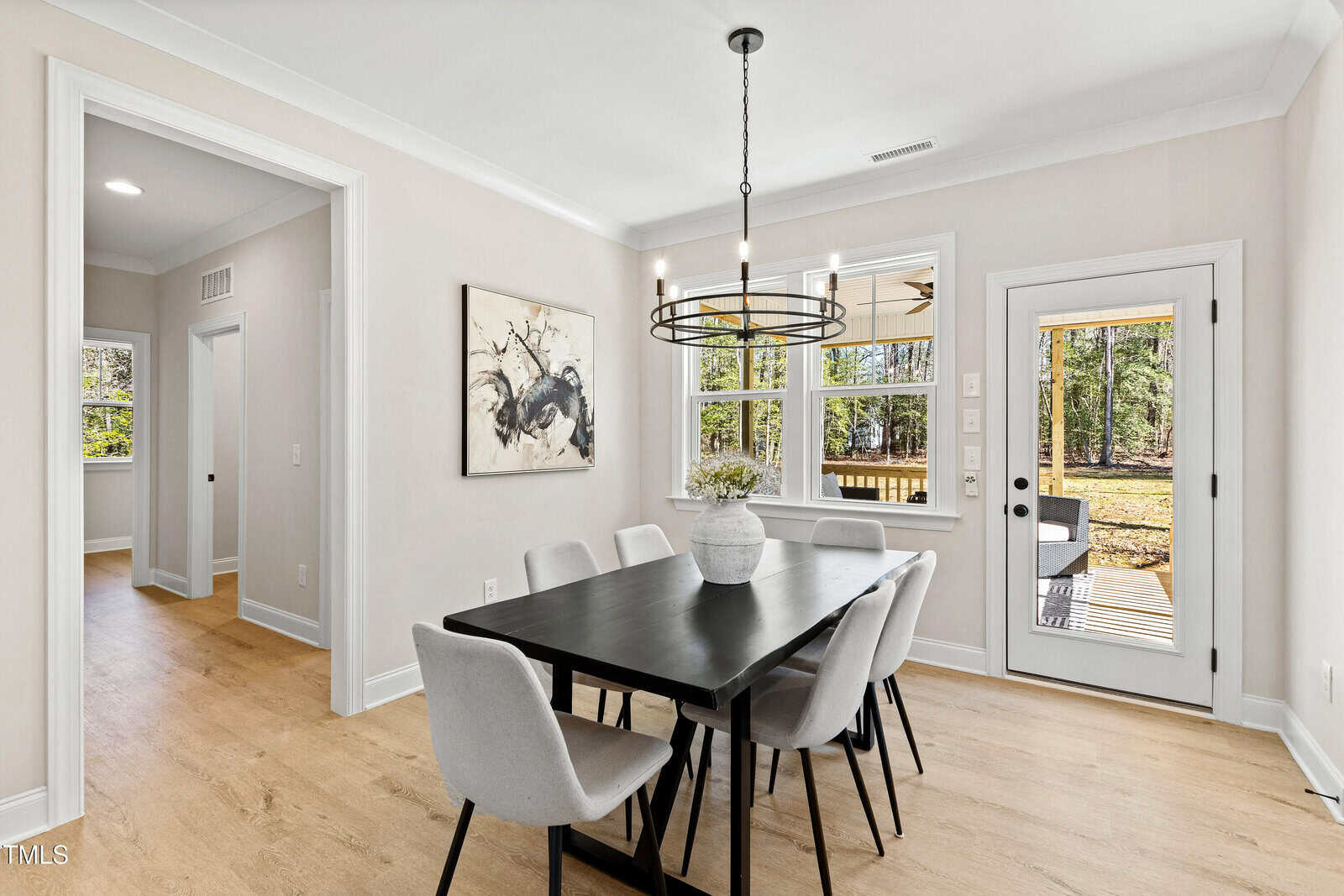 Modern dining room with a black rectangular table, six light gray chairs, a round chandelier, a potted plant centerpiece, wall art, light wood floors, and large windows and a glass door overlooking a wooded backyard.