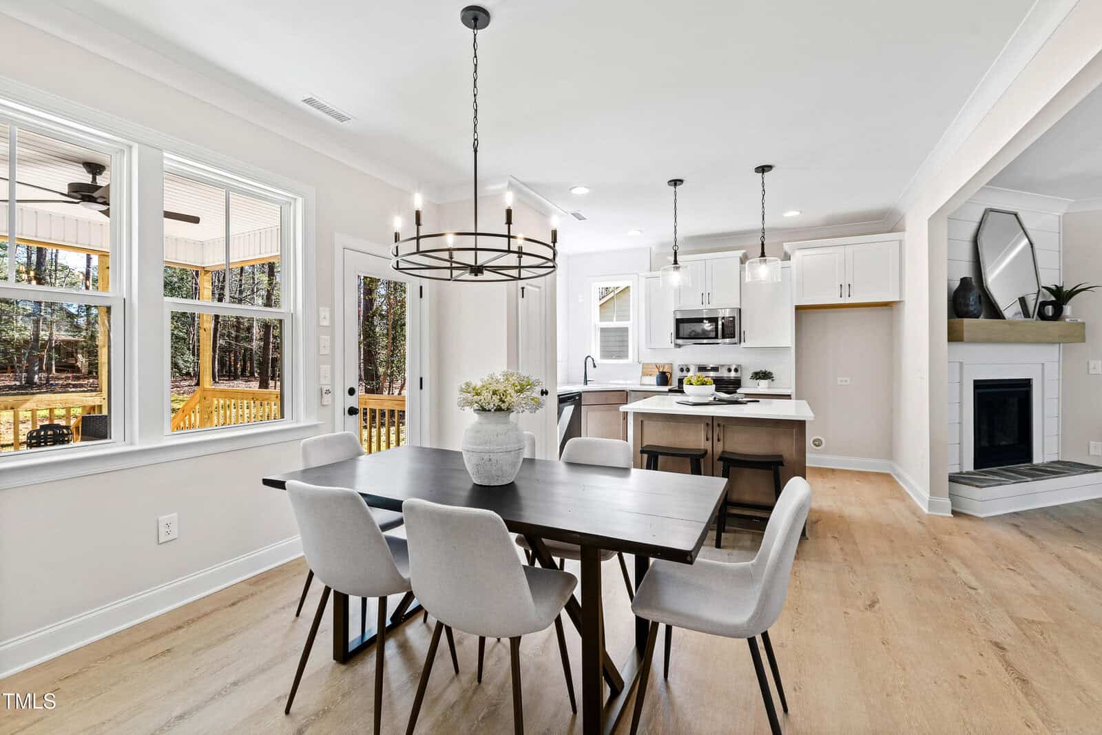 Modern dining area with a dark table, six light chairs, flower centerpiece, and a black chandelier. Open kitchen with white cabinets and bar stools. Large windows offer a view of trees and a patio. Light wood floors throughout.