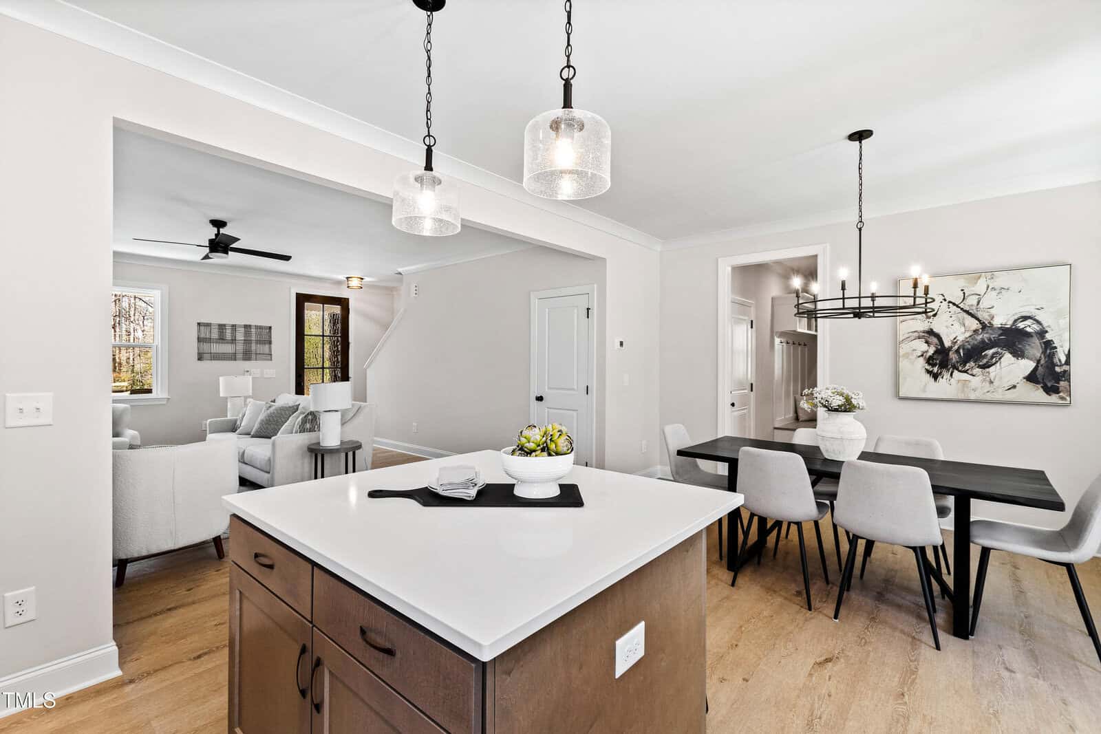 Modern open-concept dining and living area with light wood floors, white walls, a black dining table, gray chairs, and a kitchen island with pendant lights in the foreground. Contemporary art decorates the space.
