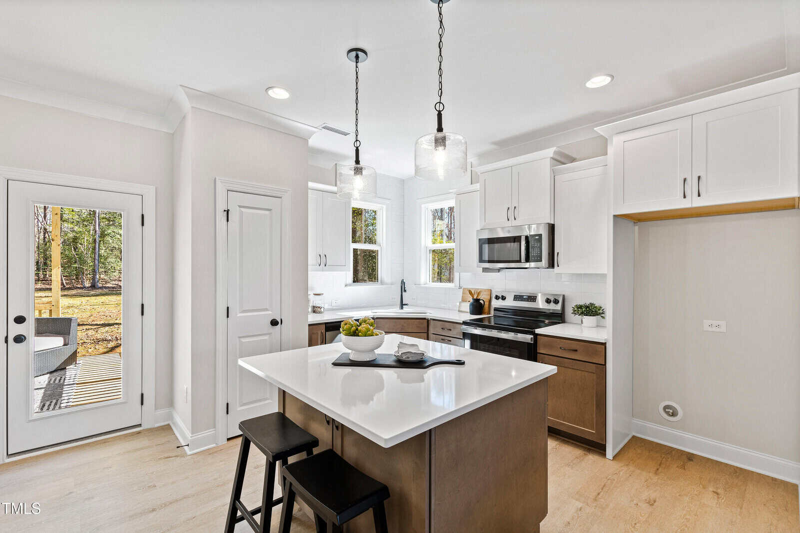 Modern kitchen with white cabinets, stainless steel appliances, a center island with two stools, pendant lights, light wood floors, and large windows letting in natural light. A door leads outside to a yard with trees.