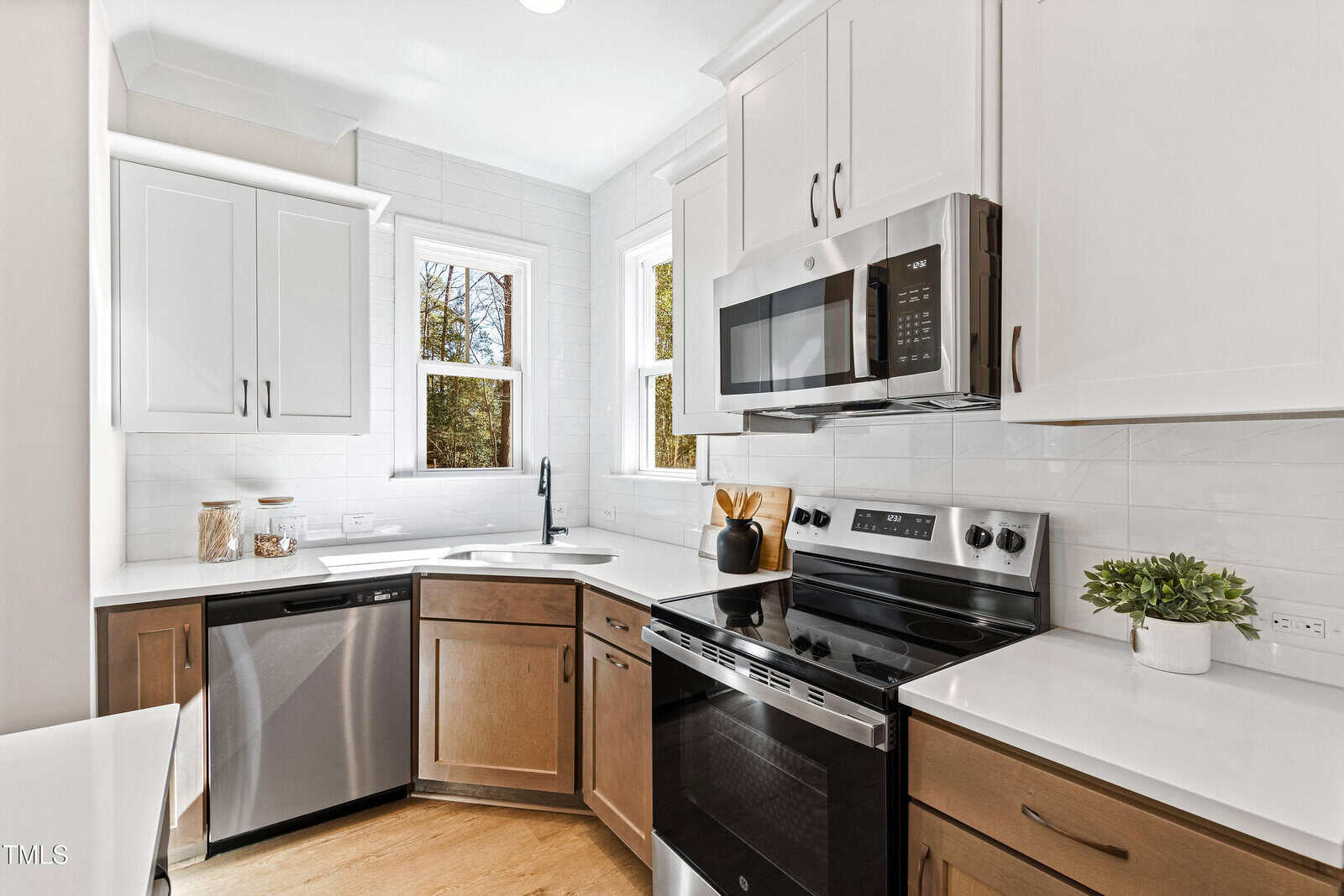 Modern kitchen with white and wood cabinets, stainless steel appliances, corner sink under two windows, white tile backsplash, and light wood flooring. A small plant and kitchen utensils decorate the countertop.