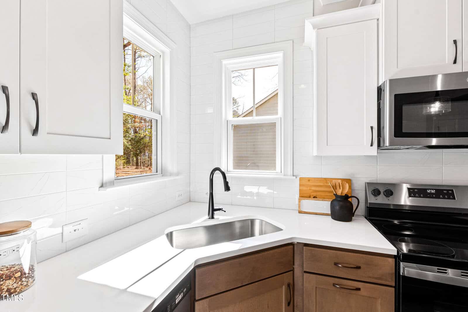Bright kitchen corner with white cabinets, a black faucet over a stainless steel sink, a cutting board and teapot on the counter, and a microwave above a black stove. Two windows let in natural light.