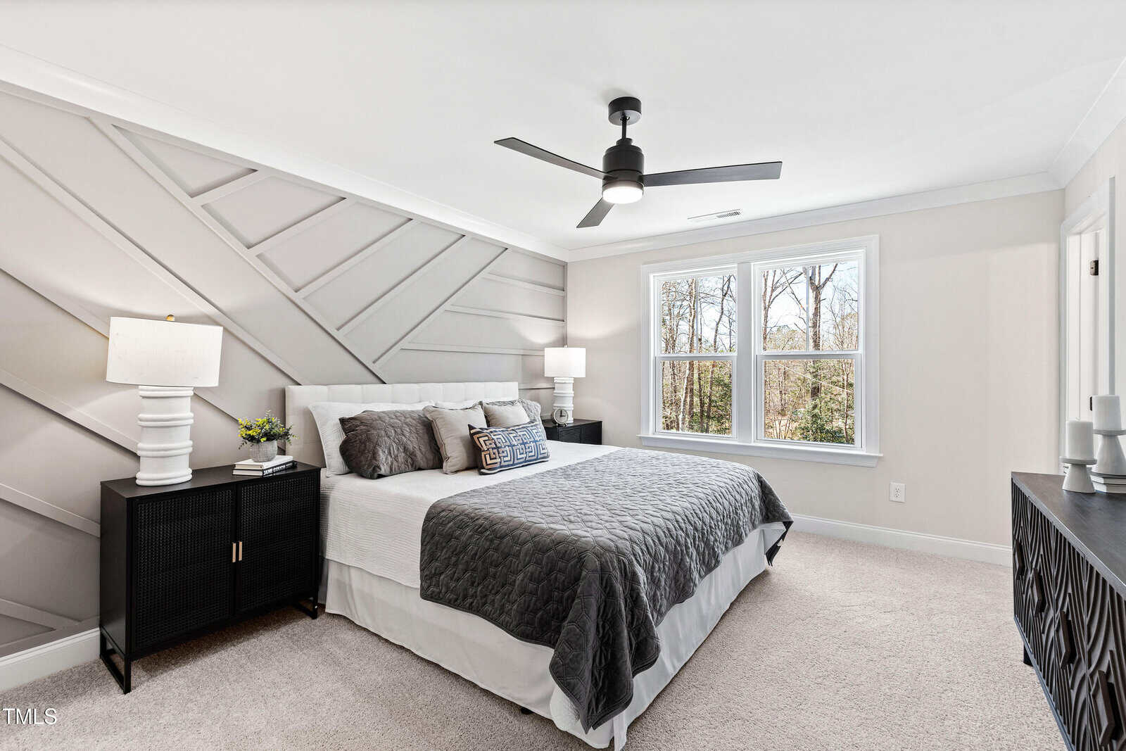 Modern bedroom with a geometric accent wall, a large bed with gray and white bedding, black nightstands with lamps, a ceiling fan, a dresser, and two windows showing trees outside.