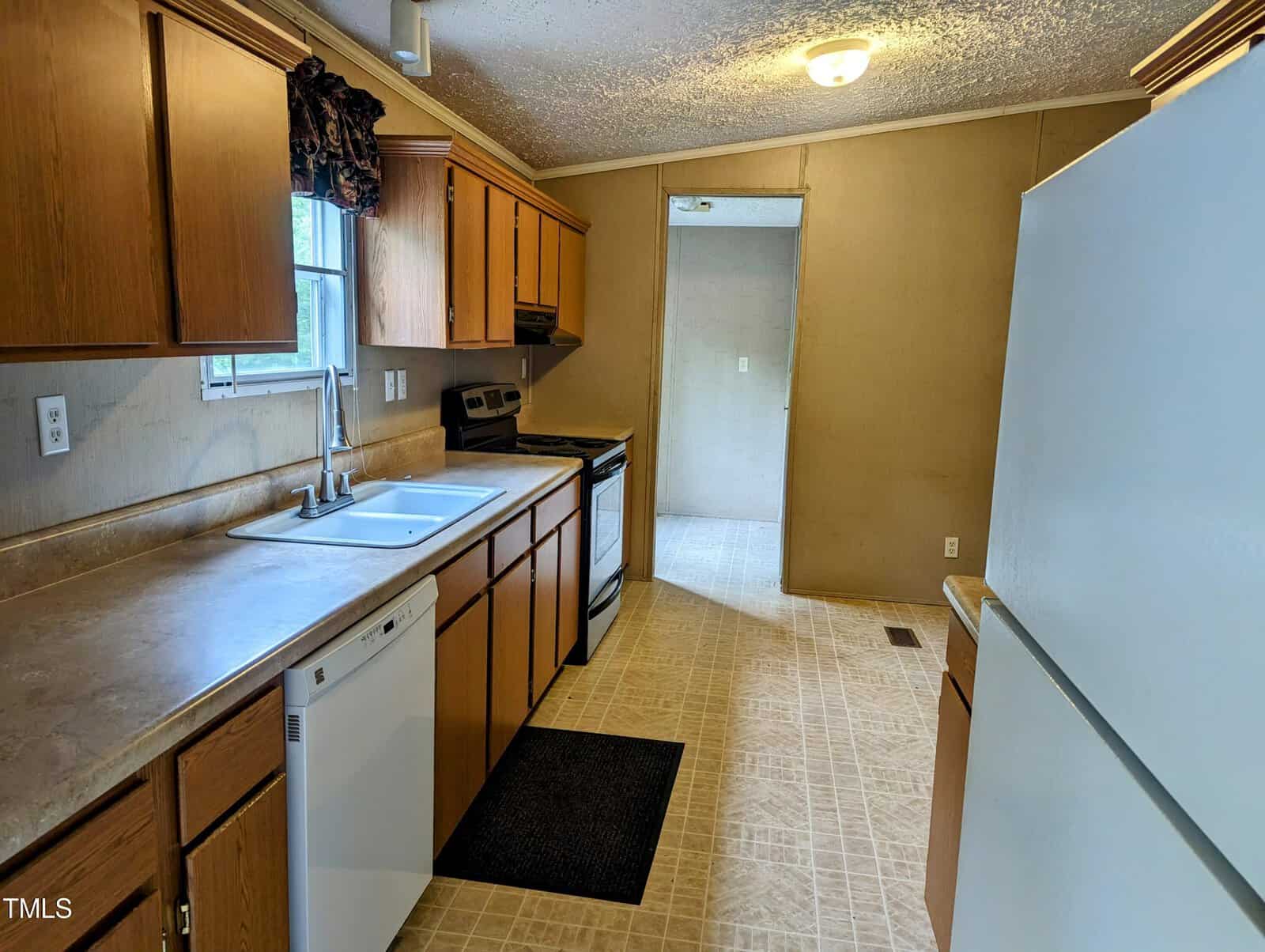 A small kitchen with wooden cabinets, marble countertops, a white double sink, white dishwasher, and a stainless-steel stove. There is a window above the sink, a white refrigerator on the right, and a door leading to another room at the far end.