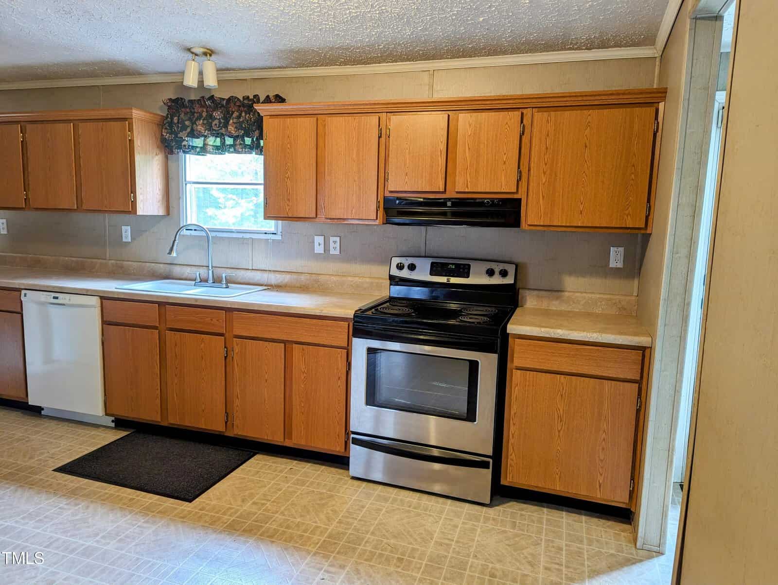 A kitchen with wooden cabinets, a stainless steel electric stove and oven, a white dishwasher, and a double sink under a window with a patterned valance. The countertops are beige, and the floor has light-colored tiles. A black rubber mat is in front of the sink.