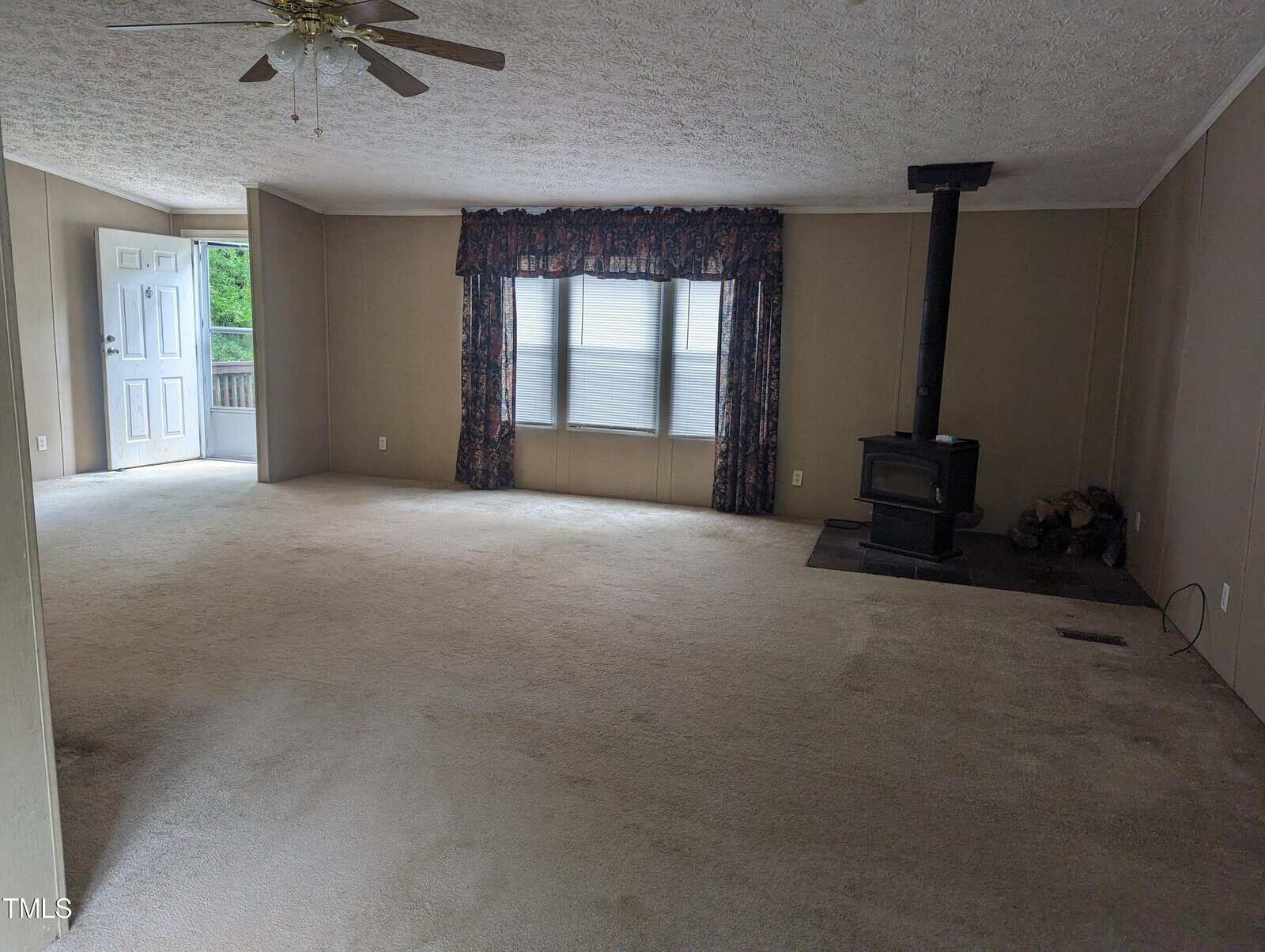 A spacious, empty living room with beige carpet and walls. There is a ceiling fan, a set of windows with floral curtains, a wood-burning stove with a small pile of firewood next to it, and an open door leading outside.