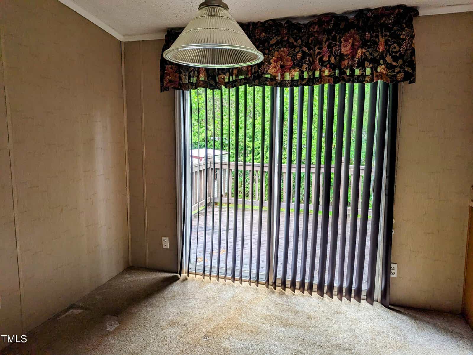 A room with beige carpet and walls features a large sliding glass door with vertical blinds leading to an outdoor wooden deck. A floral valance tops the door frame, and a hanging lamp with a wide, pleated lampshade is suspended from the ceiling.