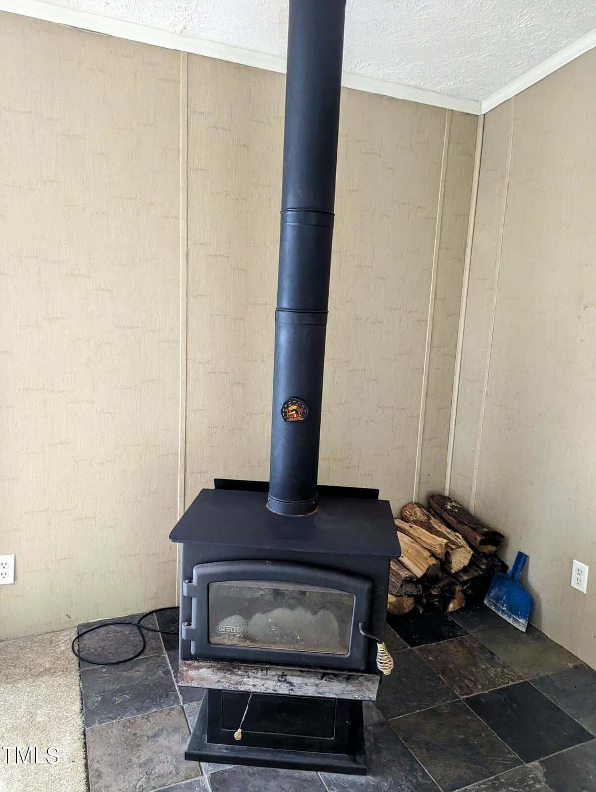 A black wood-burning stove stands against a beige wall, with a stack of firewood neatly piled in the corner on a tiled floor. An ash shovel and brush set are placed nearby on the tile. The stove has a visible stovepipe extending upwards.