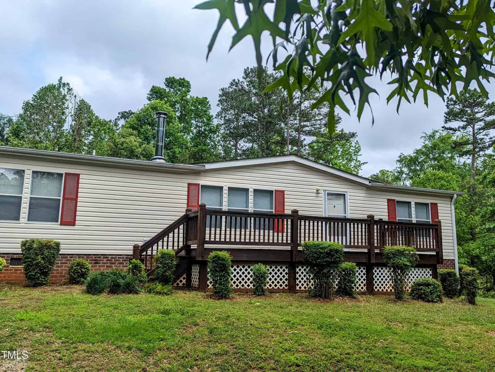 A single-story house with horizontal white siding, red shutters, and a brown wooden deck in the front. The house is surrounded by green grass, bushes, and trees, with a cloudy sky in the background. A chimney is visible on the left side of the roof.