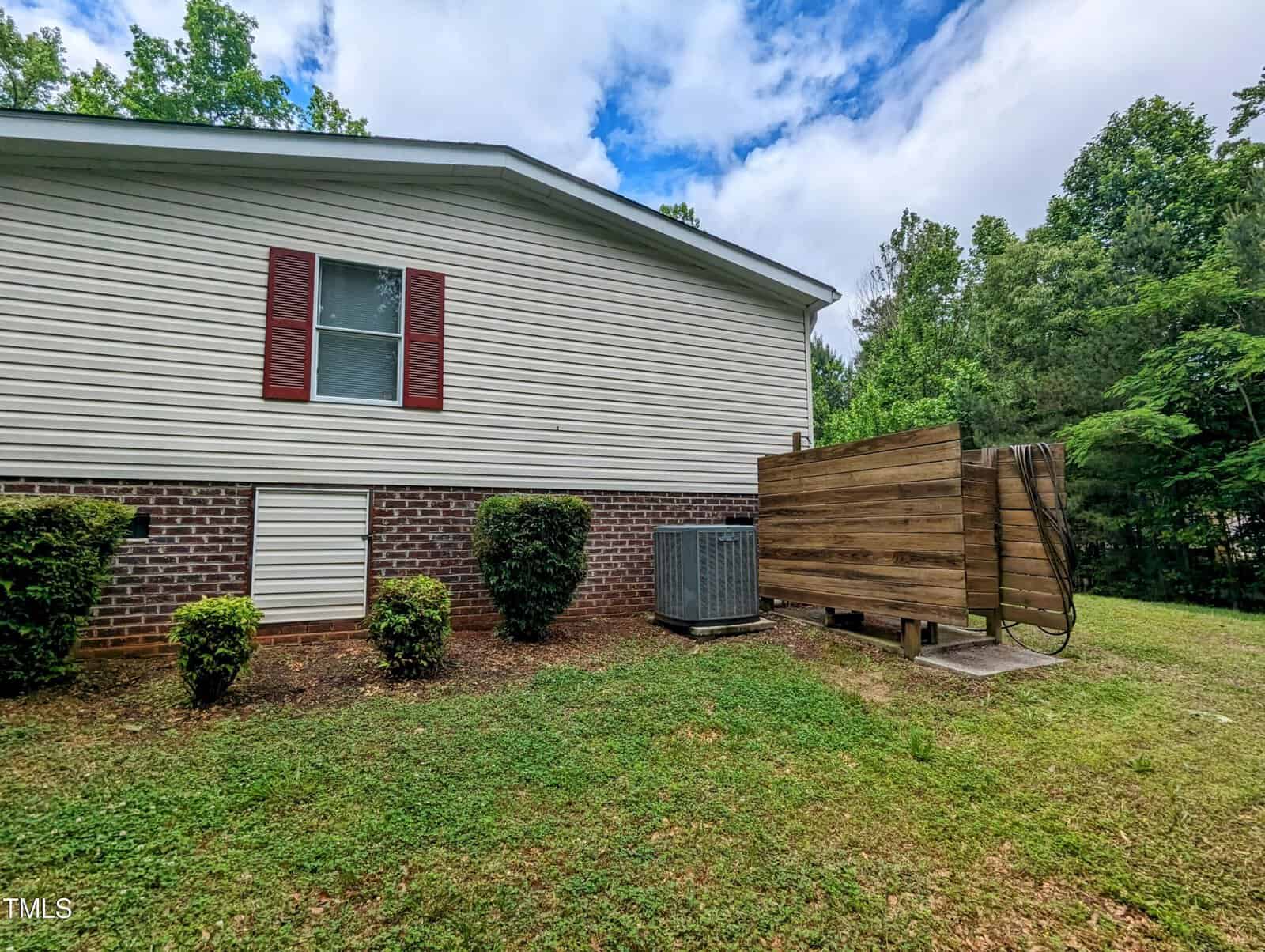 A side view of a house with a white and brick exterior, featuring a window with red shutters. An air conditioning unit and a wooden structure are positioned near the wall. Green shrubs are planted along the side, and there are trees in the background.