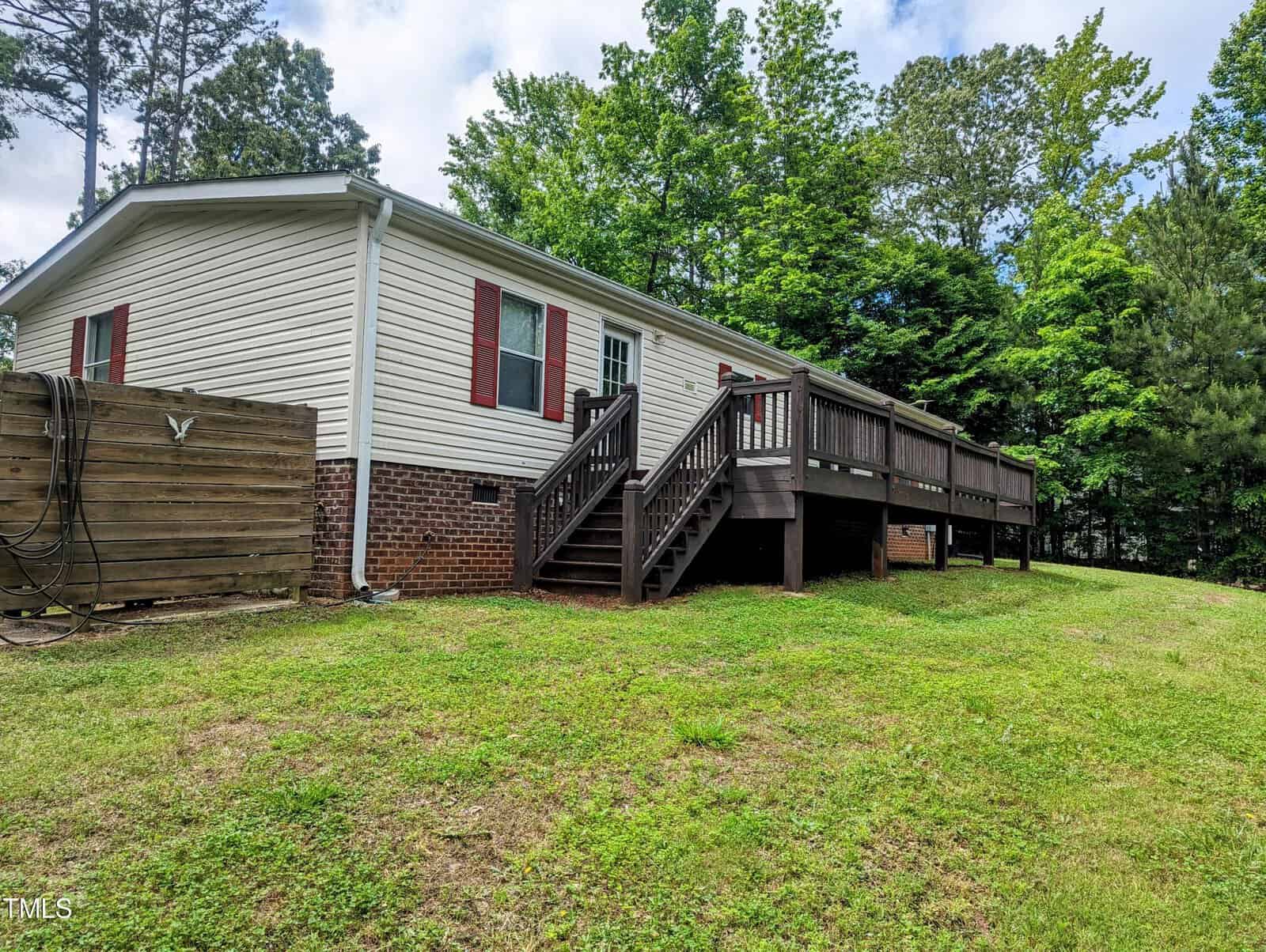 A single-story house with white siding and a brick foundation features a spacious wooden deck with stairs leading down to a grassy backyard. The house is surrounded by mature trees and greenery, providing a natural backdrop. A wooden enclosure is visible on the left side.