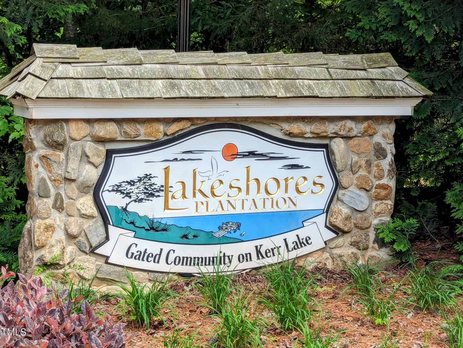 A stone sign with a wooden shingle roof reads "Lakeshores Plantation" and "Gated Community on Kerry Lake," with an illustration of a lake scene featuring trees, water, a sun, and flying birds. The sign is surrounded by greenery and some plants.