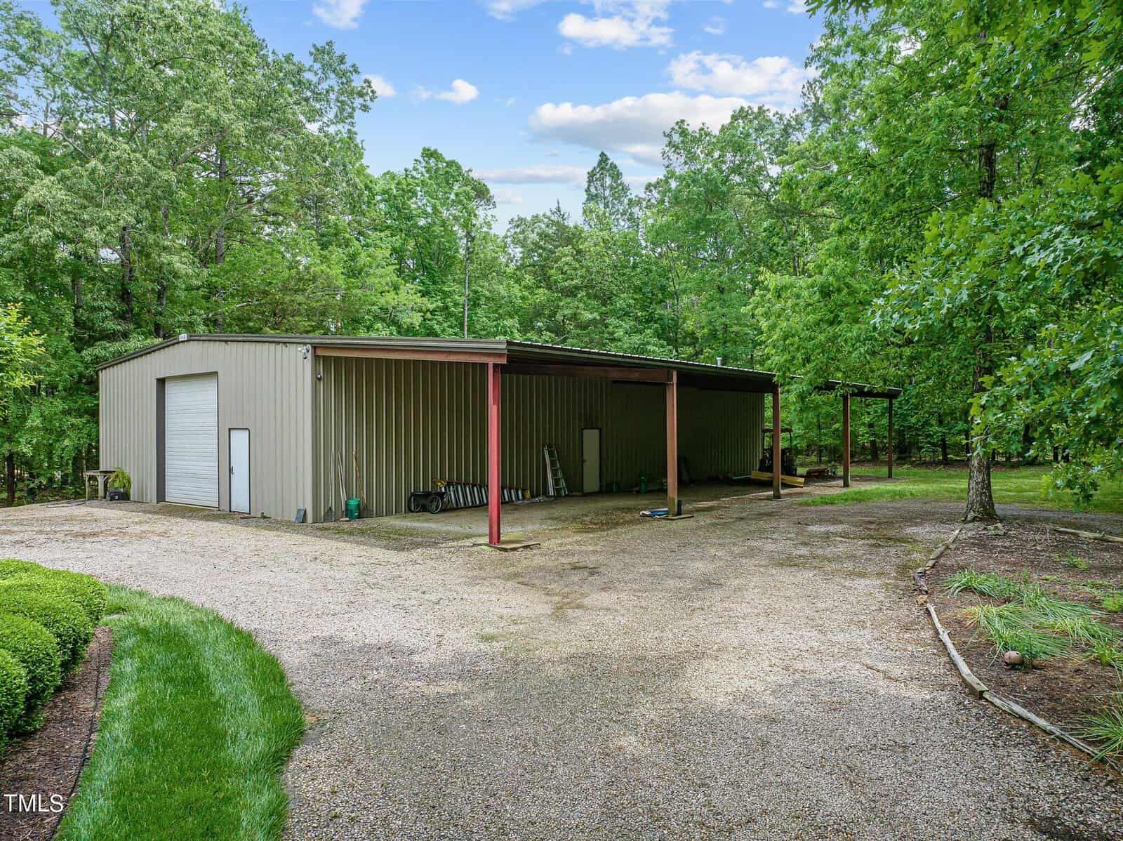 A large metal building with a sloped roof and a roll-up garage door is surrounded by trees. A covered area extends from the building over a gravel driveway. There are various items, including ladders, stored under the cover.