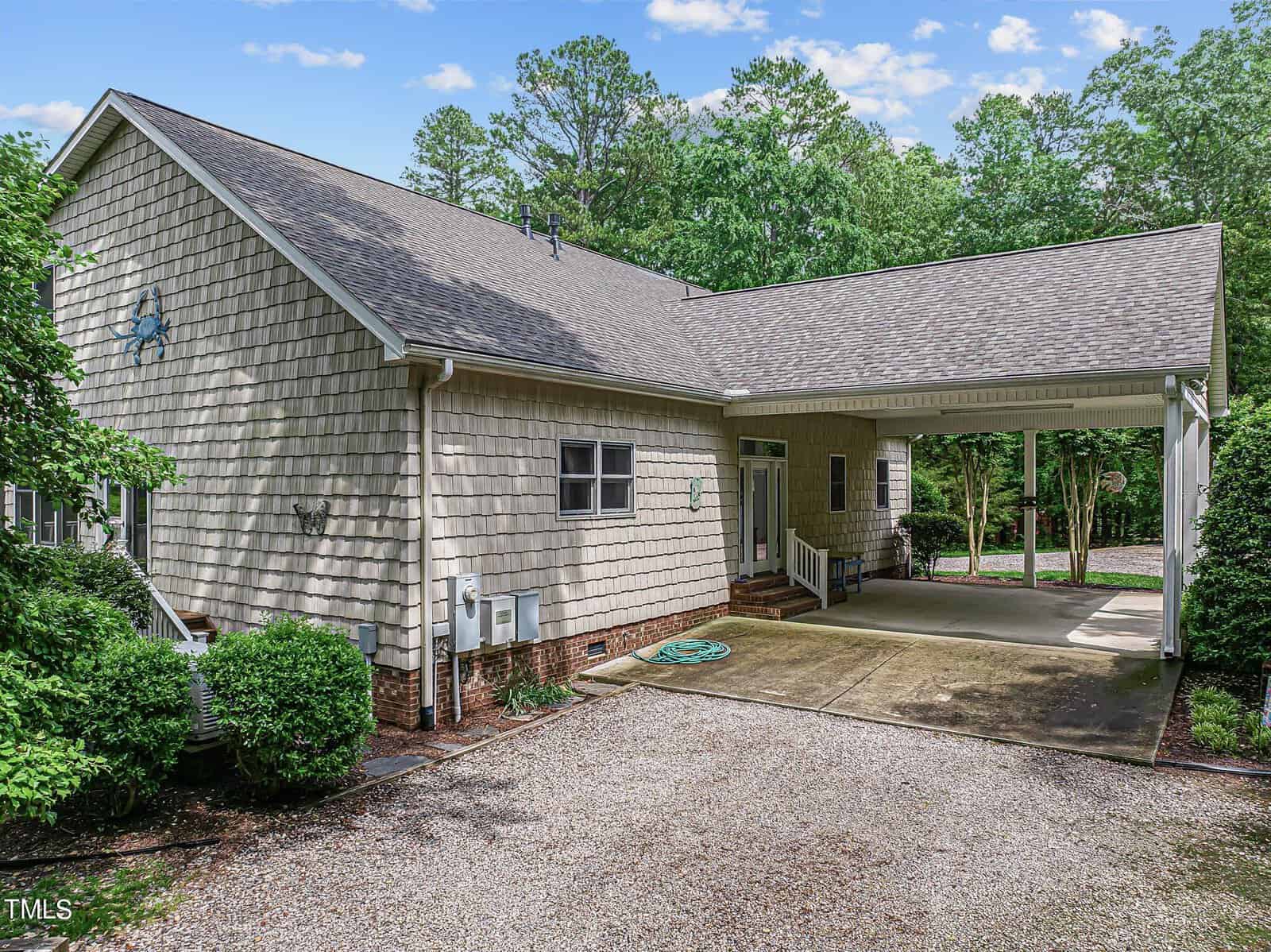 A beige two-story house with a sloped roof and a carport. The exterior has wooden shingles and a small front porch. The driveway is gravel, with trees and greenery surrounding the property. A green garden hose lies on the driveway.