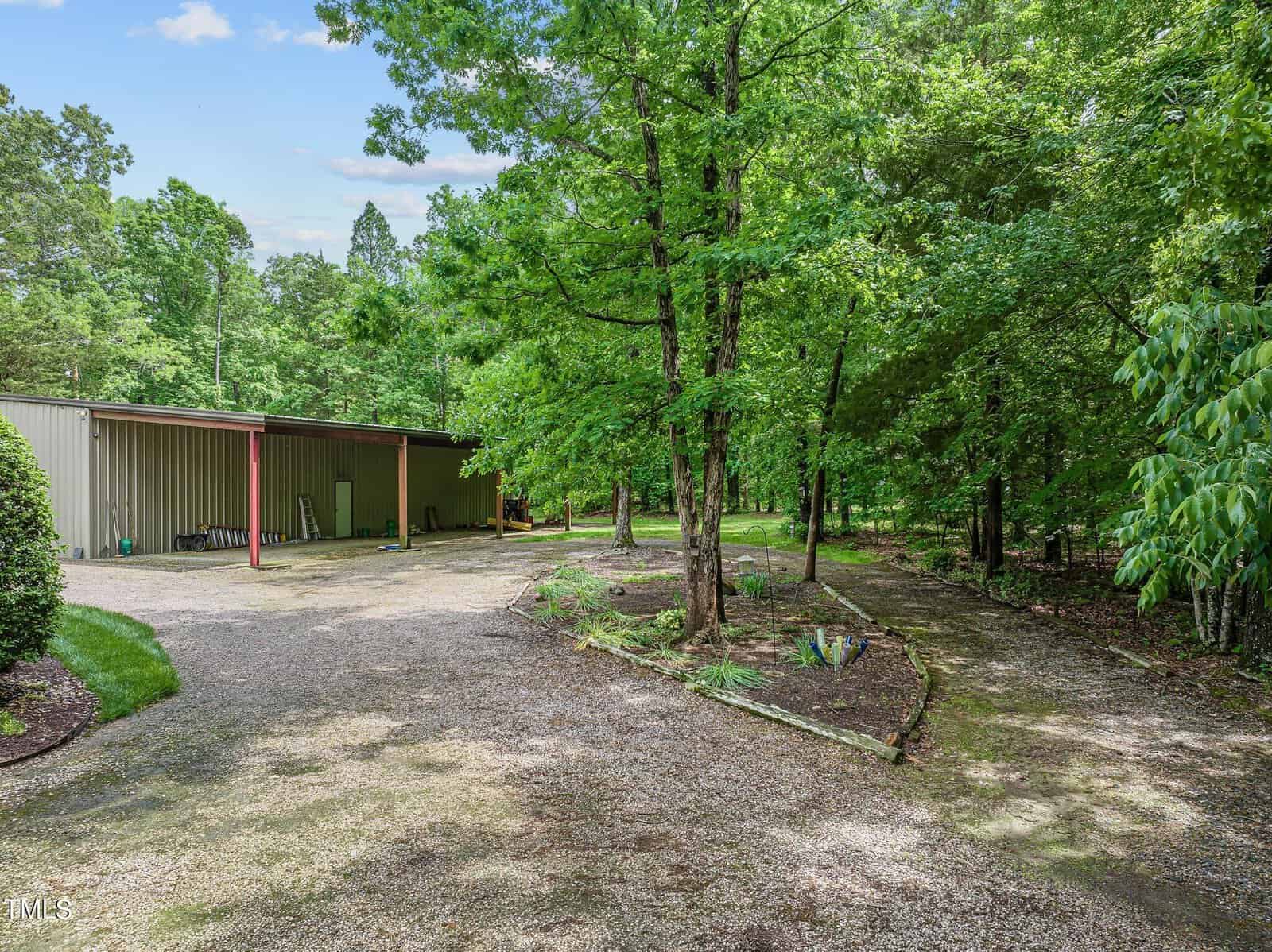 A gravel driveway leads to a metal storage building surrounded by dense green foliage and tall trees. The area is shaded, with additional gravel paths visible through the trees. The scene has a rural and natural appearance.