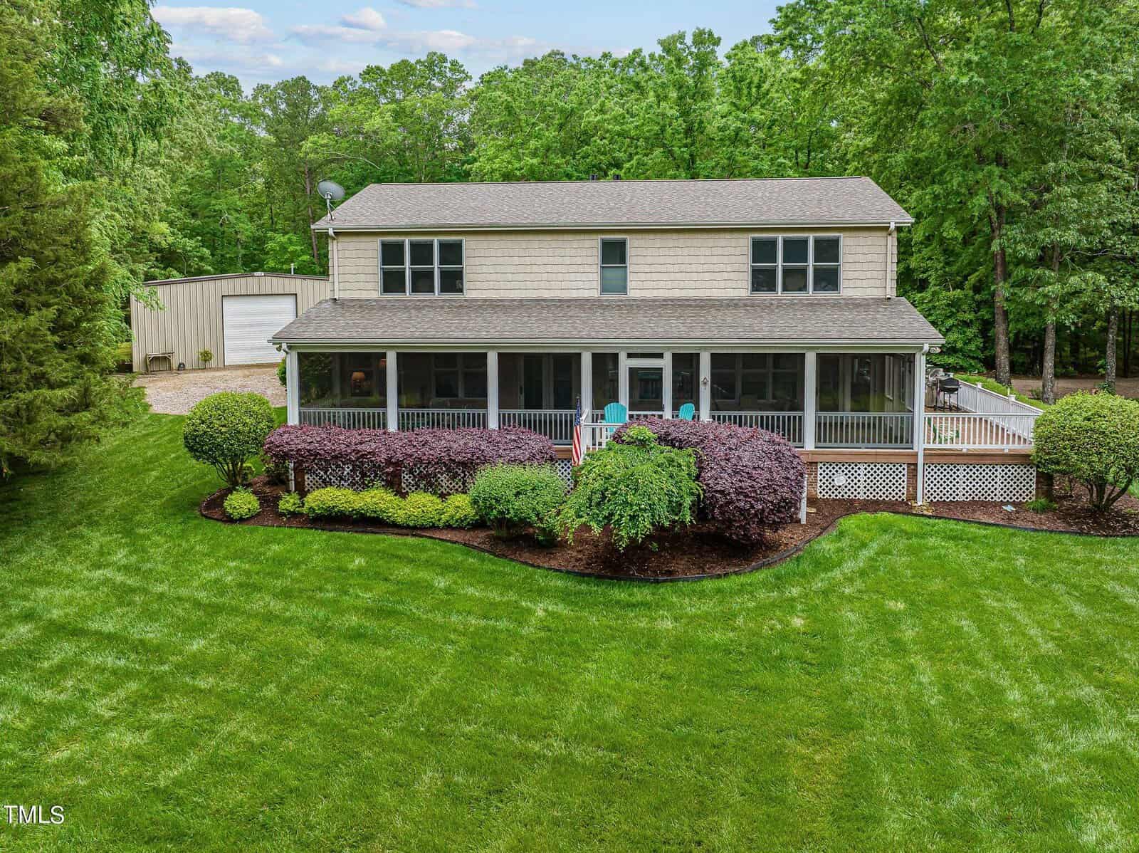 A charming two-story house at 9756 Poverty Point features a large front porch surrounded by well-maintained greenery. The freshly cut lawn and driveway to the left lead to a white Bullock garage, with trees in the background under a clear sky.