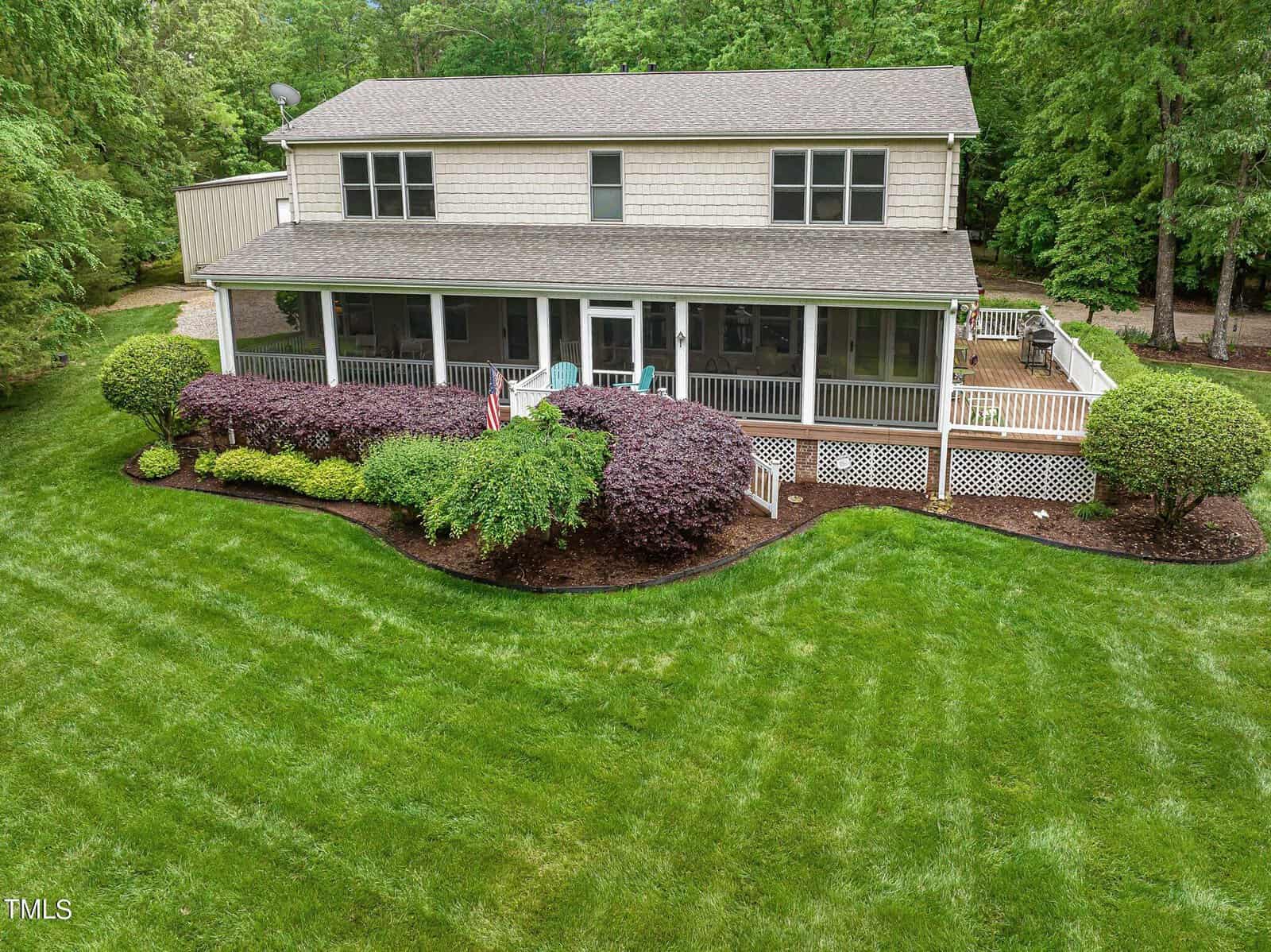 A two-story house with a large screened porch and a well-maintained lawn featuring neatly trimmed hedges. The backyard includes seating and is surrounded by a wooded area.