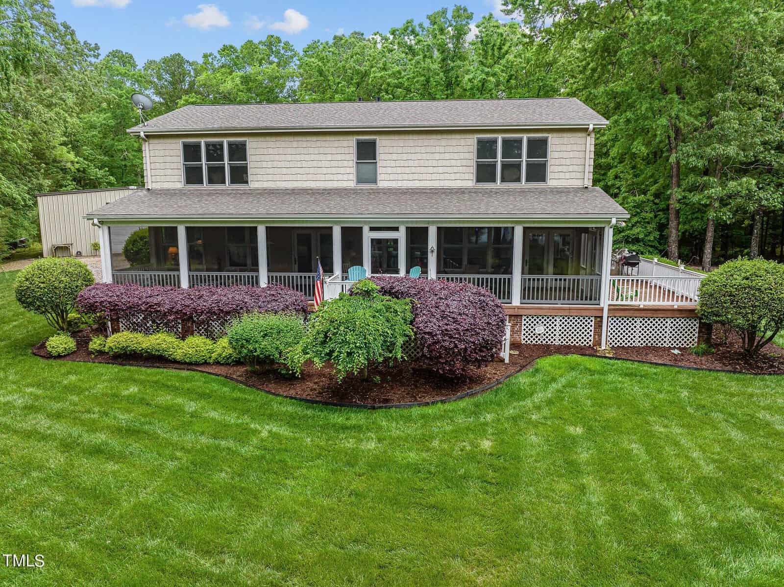 A two-story house with a large screened porch spans the front, surrounded by neatly trimmed bushes and a well-maintained lawn. A flag is mounted near the porch entrance. Tall trees line the background.