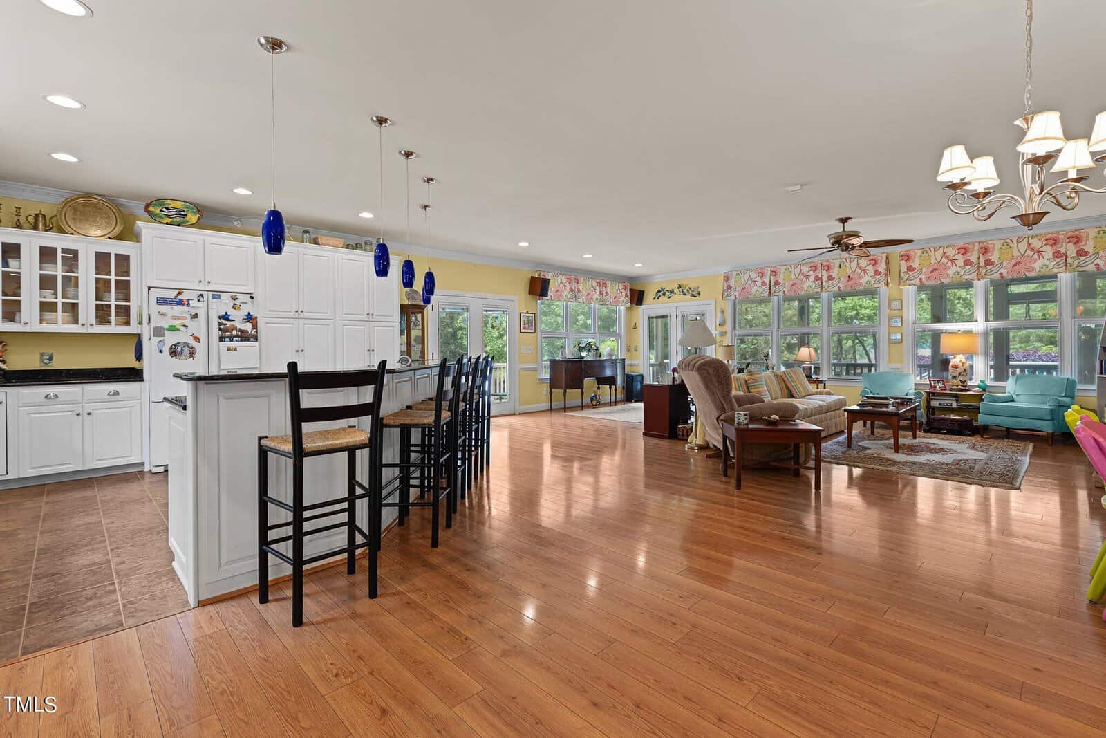 Open-concept living space featuring a kitchen with white cabinetry and blue pendant lights, a breakfast bar with tall chairs, and a living area with hardwood floors, floral-patterned window valances, and a variety of seating options.