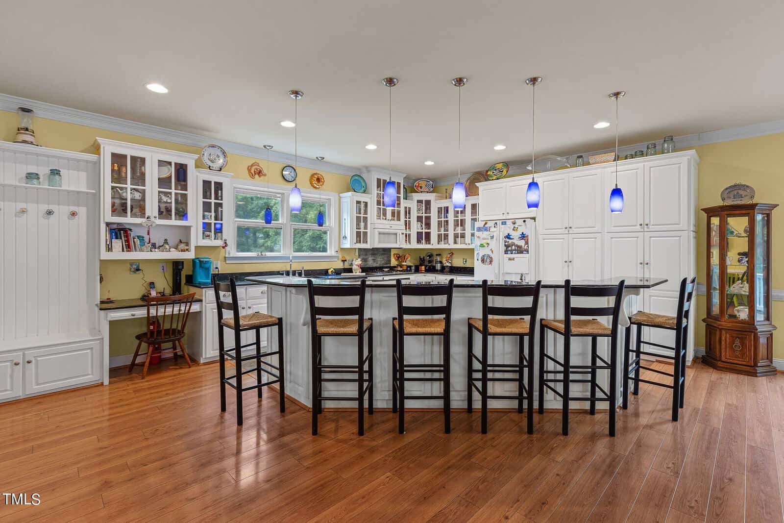 A spacious kitchen with wooden flooring features a long counter with seven tall chairs. White cabinets, blue pendant lights, and a variety of decor items are seen. A desk with a chair and a glass display cabinet are located on the left side.