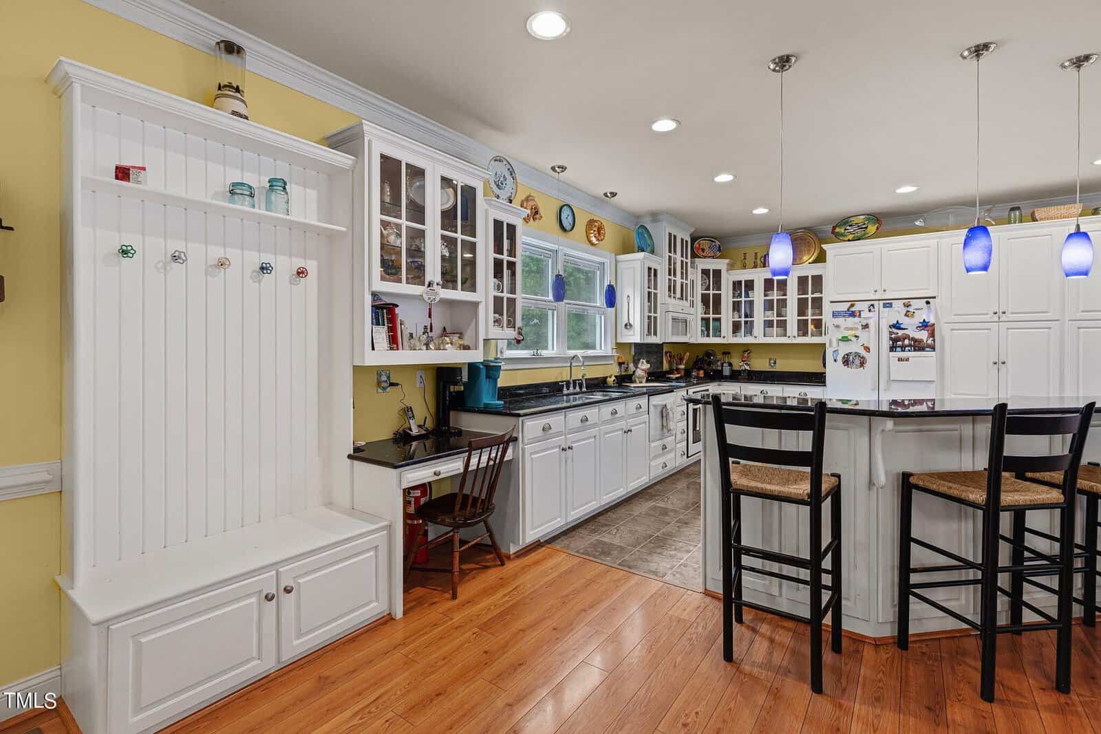 A spacious kitchen features white cabinetry, glass-front cupboards, and a black countertop. It has a central island with high chairs and pendant lights. Wooden flooring contrasts with the yellow walls and recessed ceiling lights provide illumination.