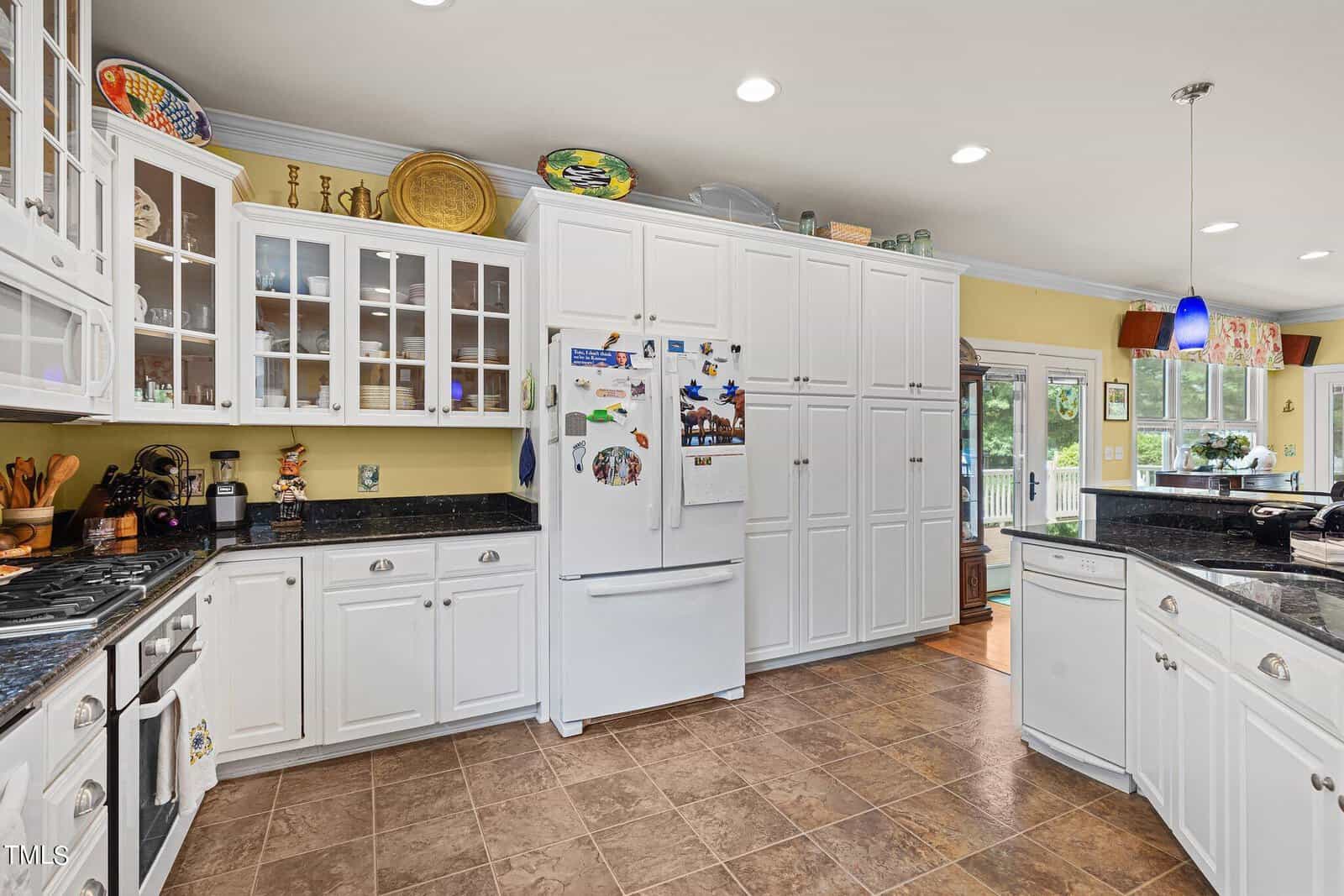 A spacious kitchen featuring white cabinetry and glass-front cabinets. A white refrigerator is adorned with various magnets. The countertops are black, and a window provides natural light. Blue pendant lights hang over the island.