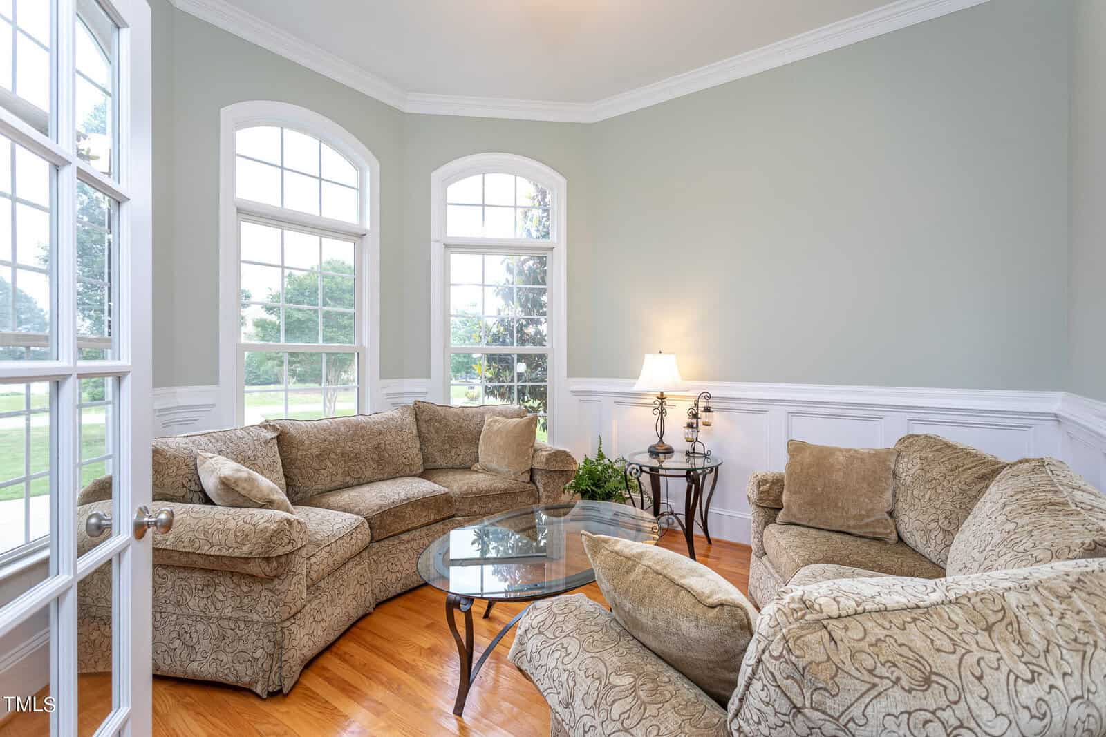 A cozy living room with a large patterned sectional sofa, a round glass coffee table, and decorative pillows. The space is brightened by three large windows and complemented by a table lamp in the corner. The walls are painted light green.