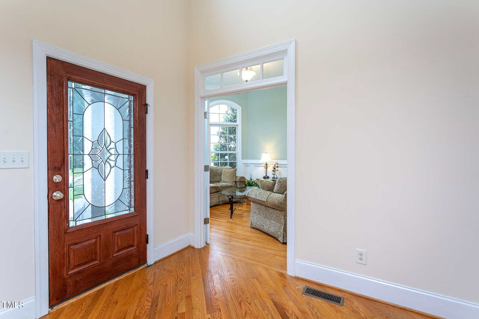 A brightly lit entryway with a wooden front door featuring decorative glass. Adjacent is a doorway leading to a cozy living room with large windows, cream walls, and a sectional sofa. The floor is polished hardwood.