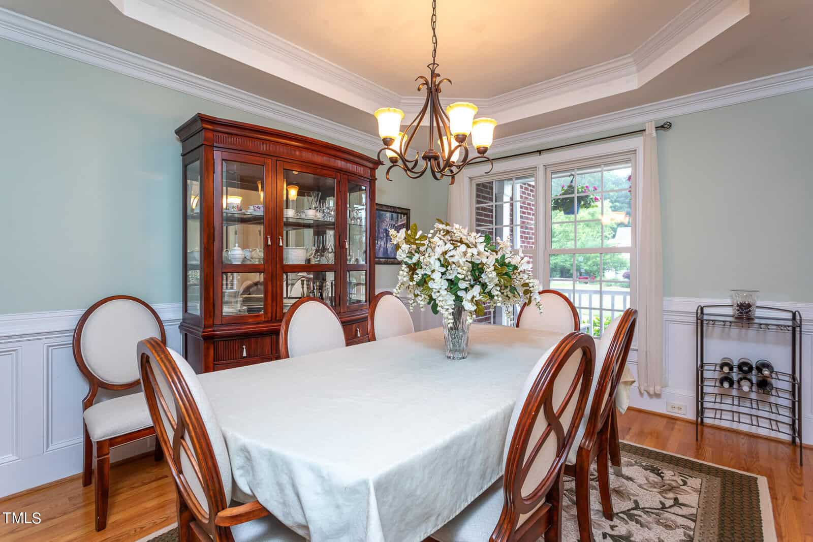 A dining room features a wooden dining table with eight upholstered chairs, a floral centerpiece, a glass-front cabinet displaying dishes, and a chandelier overhead. Large windows let in natural light, and a wine rack is in the corner.
