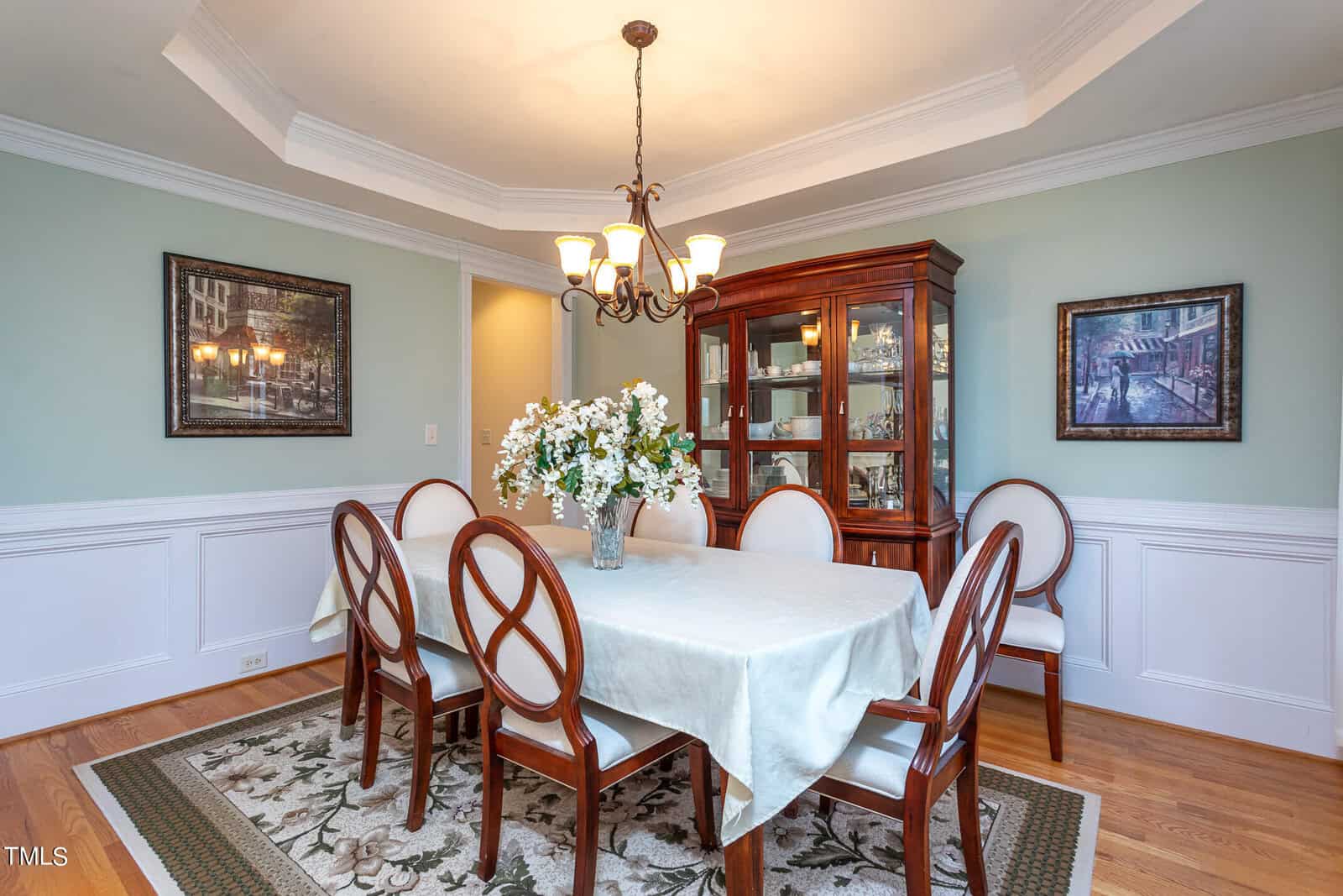 A dining room with a table covered by a light blue tablecloth and surrounded by six wooden chairs. A chandelier hangs above the table, which displays a vase of flowers. A wooden cabinet and two framed artworks adorn the pale green walls.