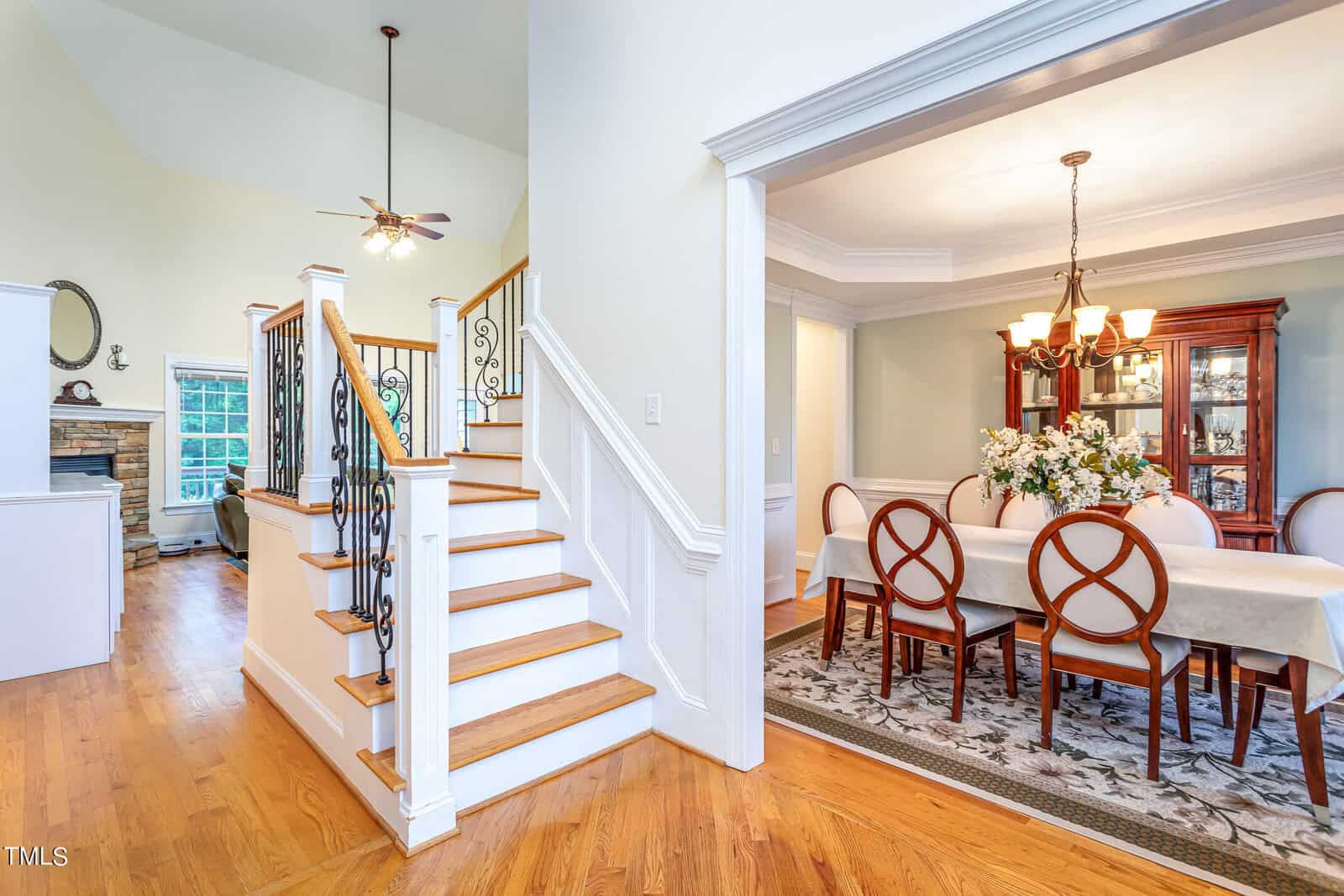 A contemporary home interior with wooden floors features a staircase with wrought iron railings. To the right, a dining room with a table set for eight, a chandelier, and a cabinet. A living area with a stone fireplace and ceiling fan is visible.