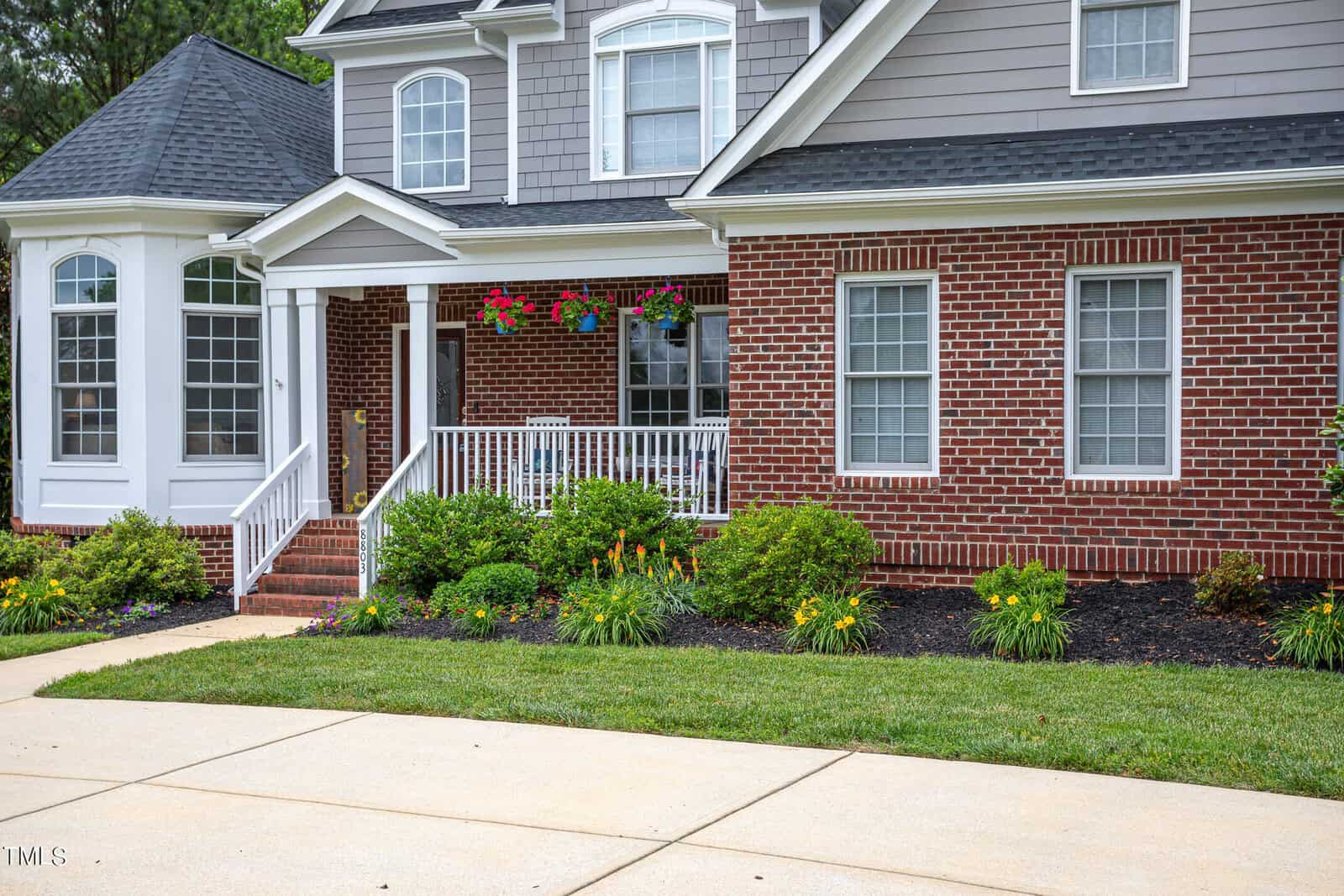 A two-story house with a brick facade, white-trimmed windows, and a covered porch. It features hanging flower baskets, a neatly landscaped garden with green shrubs, and a paved driveway.