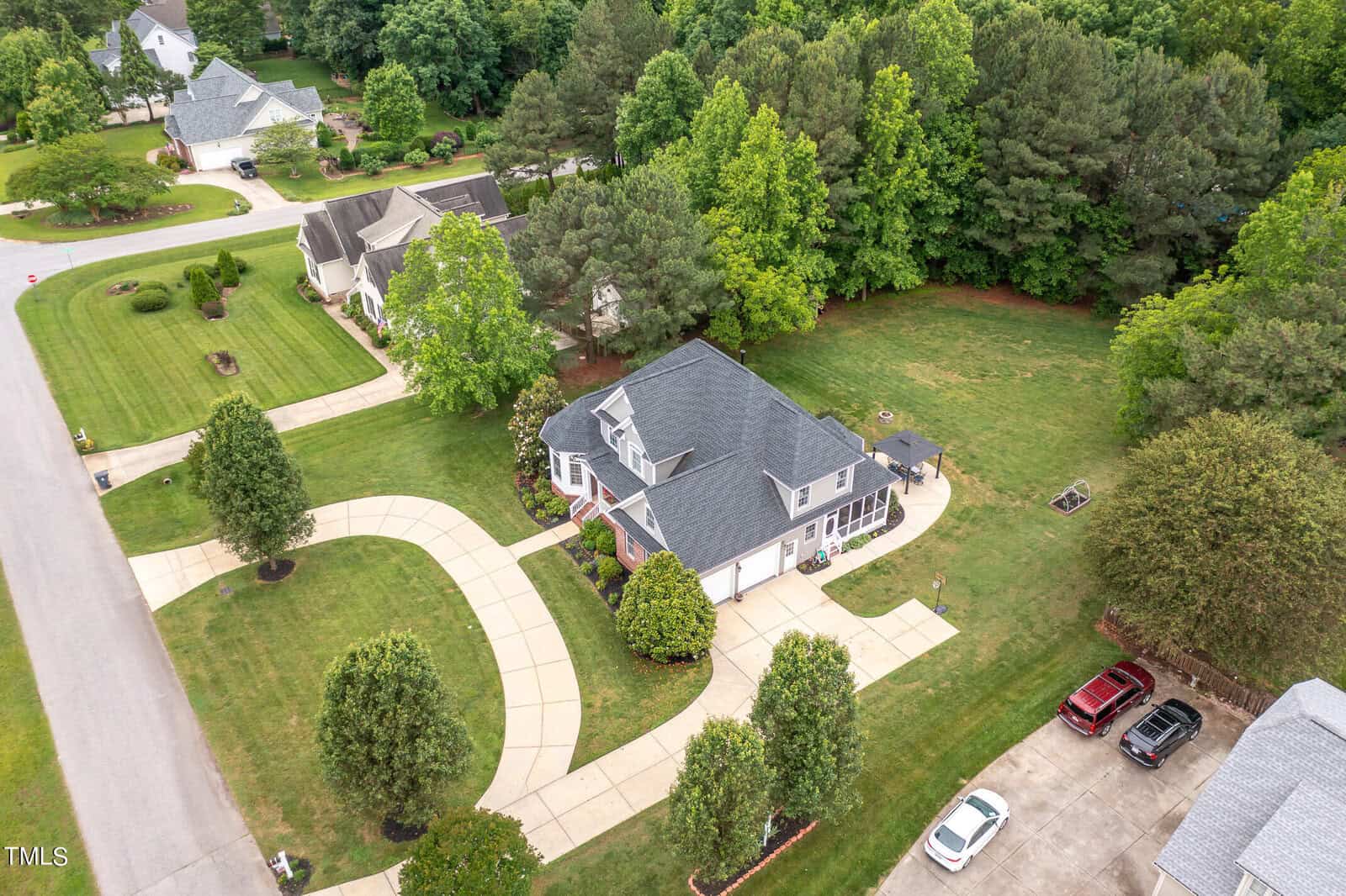 Aerial view of a suburban neighborhood featuring a large house with a gray roof and white exterior. It has a circular driveway, lush green lawns, and is surrounded by trees. Adjacent houses and a few parked cars are visible.