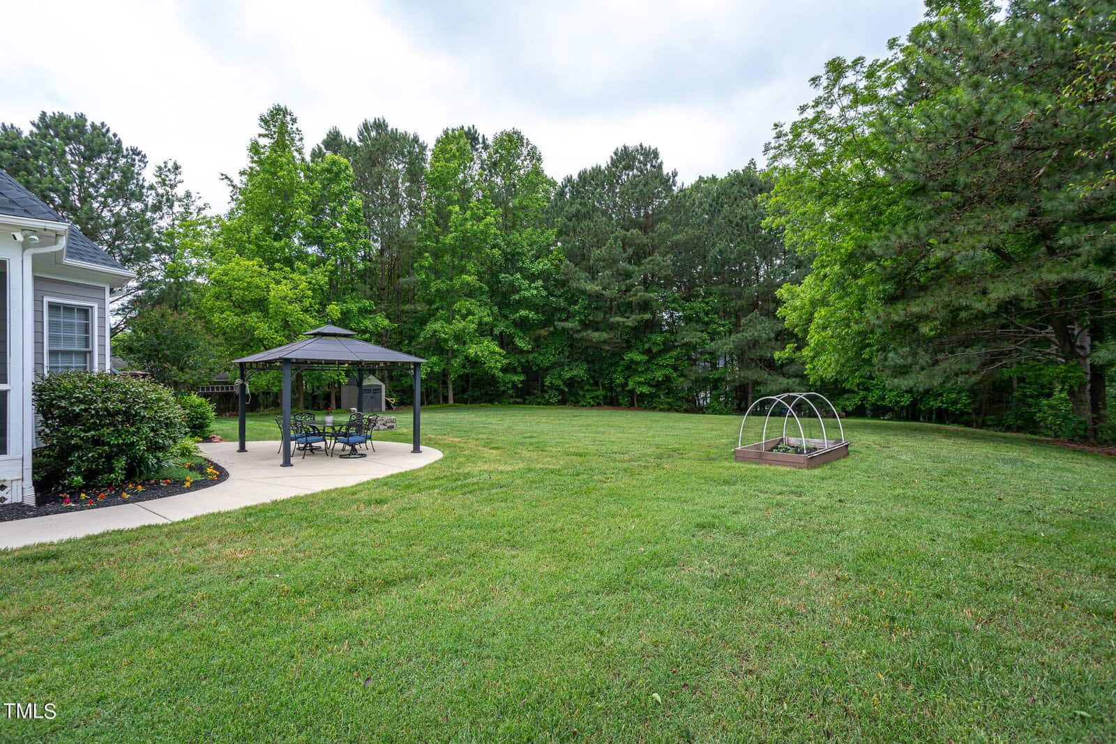 A spacious backyard with a gazebo on a concrete patio, a table and chairs underneath. There's a small garden bed with a transparent cover nearby. The area is surrounded by lush green grass and tall trees.