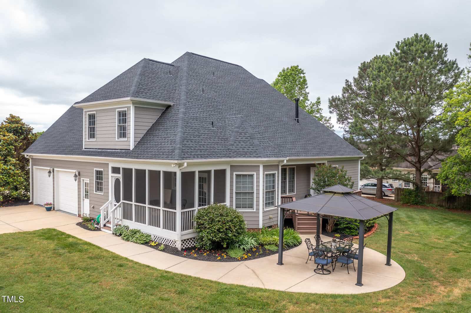 A large, two-story house with a gray roof and beige siding features a wraparound porch. A paved patio area includes a gazebo with a table and chairs. The yard is well-maintained with shrubs and a lush green lawn.
