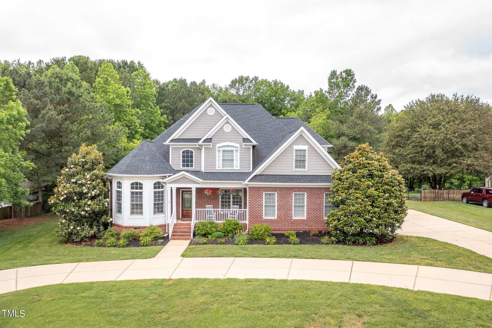 Located on 8803 Sunflower Meadows Lane in Wake Forest, this two-story suburban house with gray siding and brick accents features a gabled roof and white columns on the porch. It is surrounded by a lush green lawn, trees, and a curved driveway.