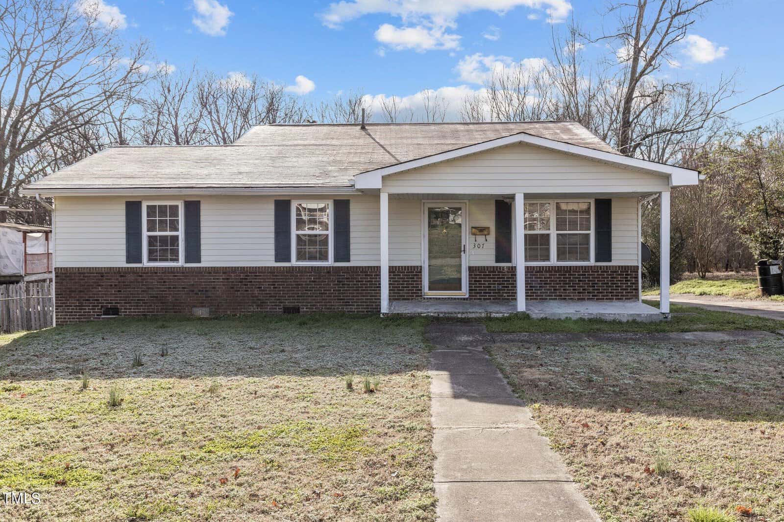 A small, single-story house at 307 Cherry Street with a brick and white exterior, featuring a front porch and dark shutters. This Oxford home is surrounded by a grassy yard and bare trees under a clear blue sky with a few clouds. A concrete walkway leads to the entrance.